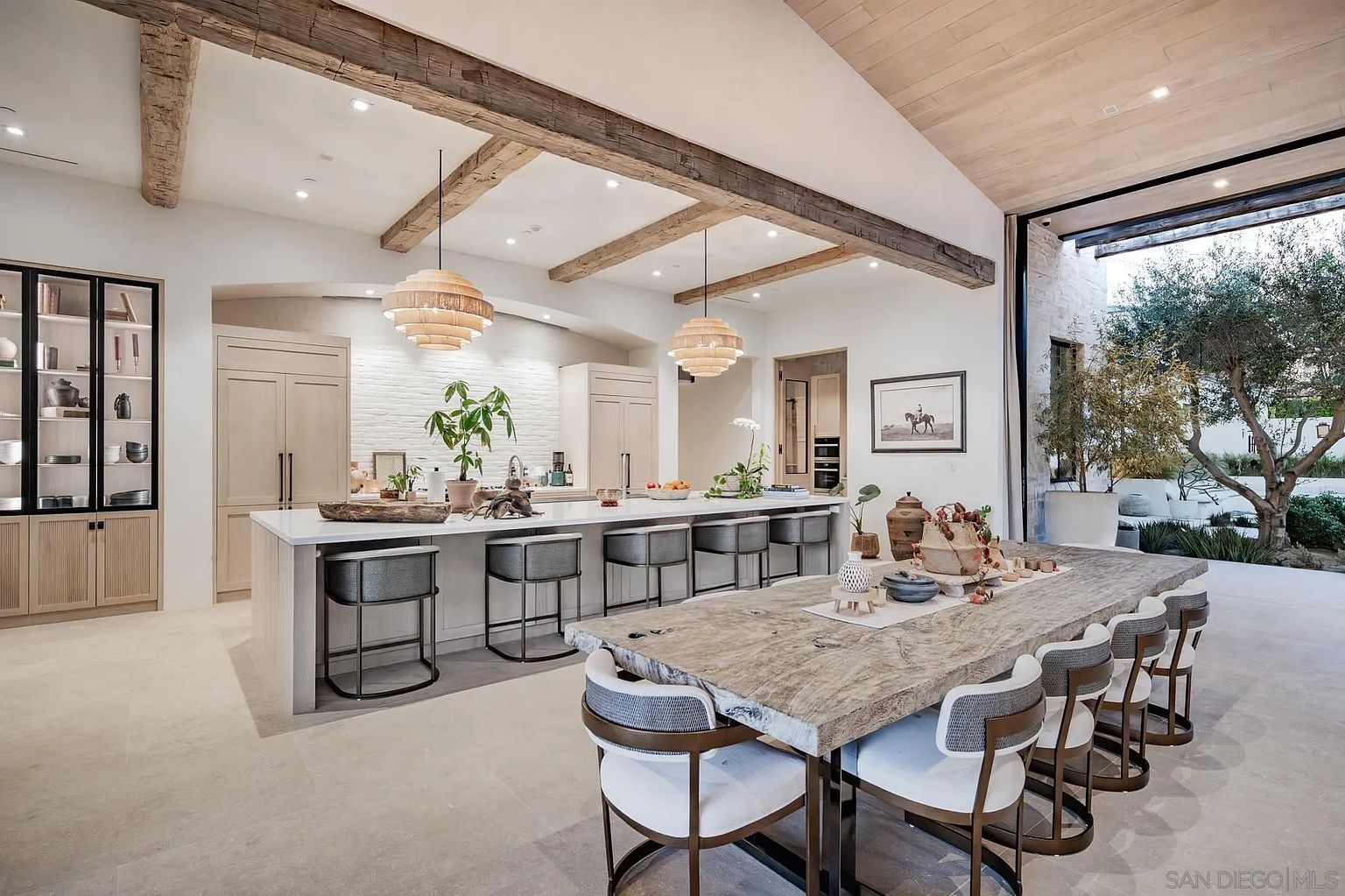 This expansive, open-concept kitchen and dining area features a large central island with bar seating and a rustic, live-edge wood dining table. The space is defined by exposed wooden ceiling beams, light-toned cabinetry, and a white brick backsplash, creating a warm, modern farmhouse aesthetic. A seamless transition to the outdoors is provided by a large, retractable glass wall that opens to a patio with a mature tree.
