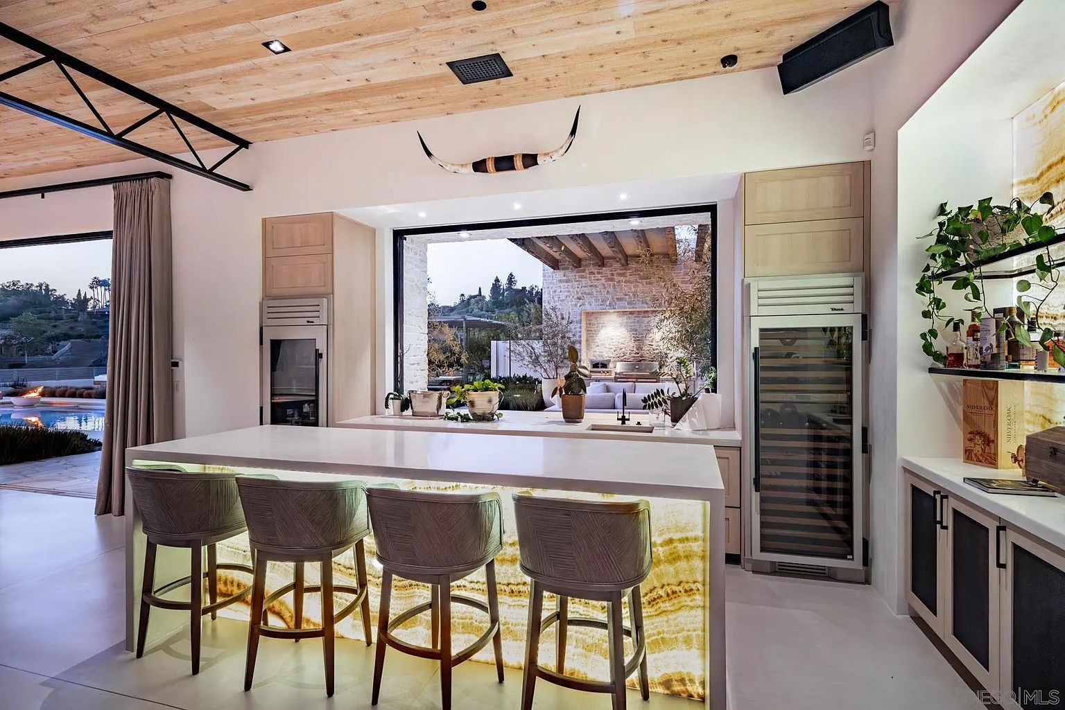 This sophisticated kitchen features a large central island with a backlit stone base and four bar stools, serving as the focal point of the room. The space is defined by light wood ceilings, a large window looking out to an outdoor patio, and integrated high-end appliances including a wine refrigerator. A decorative longhorn mount hangs above the window, adding a rustic touch to the otherwise modern and airy design.