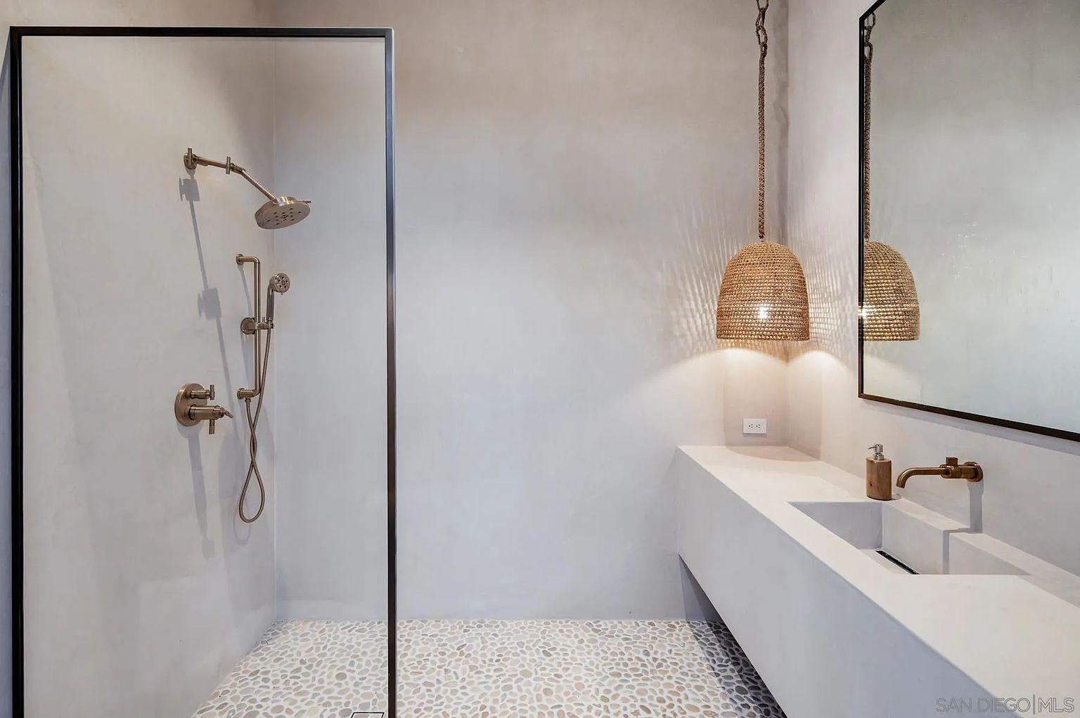 This modern bathroom features a minimalist aesthetic with smooth, light-toned walls and a pebble-tiled shower floor. A glass-enclosed shower area is equipped with bronze-toned fixtures, while a floating vanity with a built-in sink sits beneath a large mirror. A textured, woven pendant light hangs from the ceiling, adding a warm, organic touch to the clean, contemporary space.