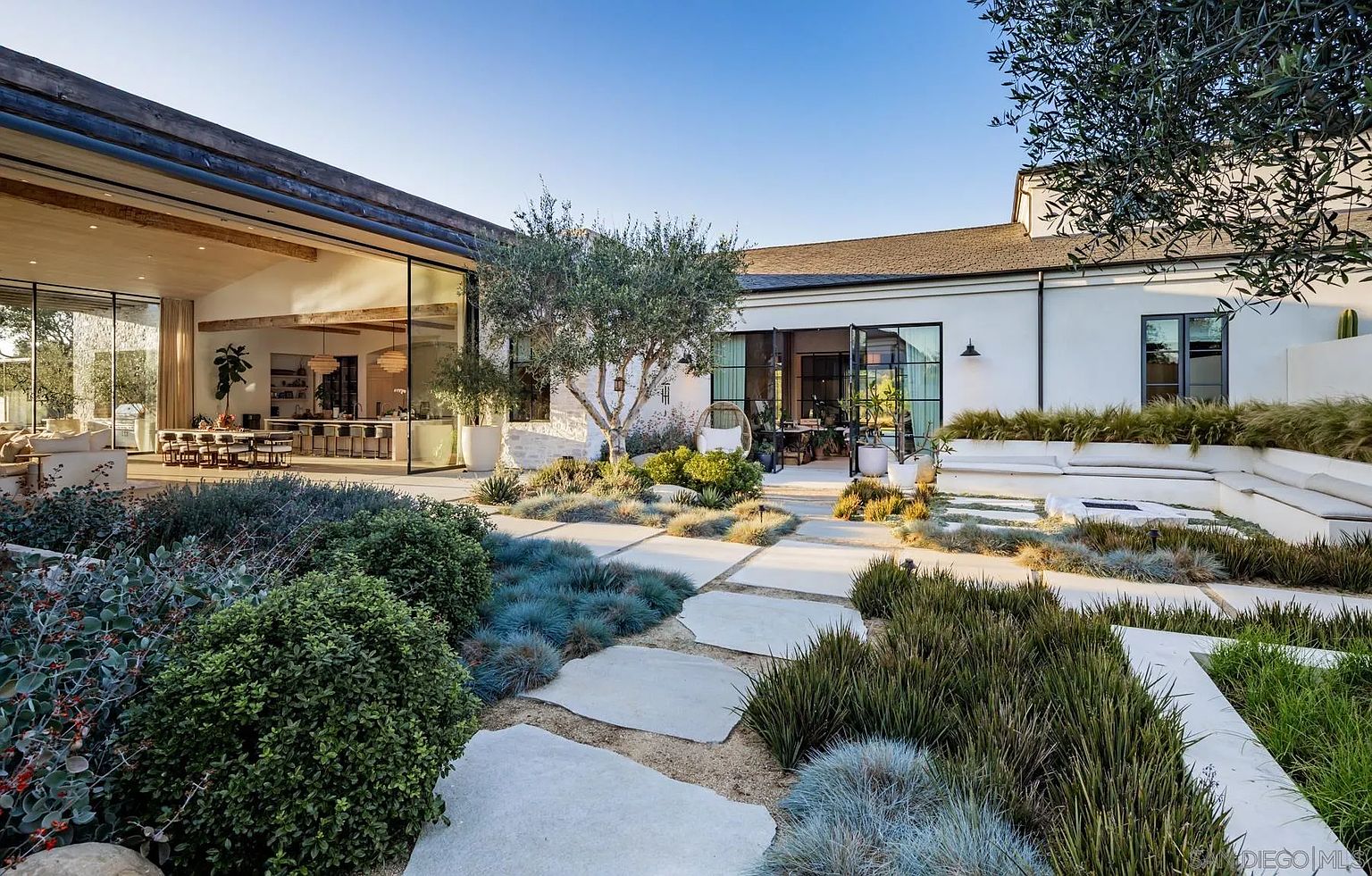 This expansive outdoor living space features a modern, minimalist garden with large, irregular stone pavers set into light-colored gravel. The scene highlights a seamless transition between the indoor living area and the exterior, facilitated by large glass walls that open to a dining space and kitchen. A built-in tiered seating area surrounds a fire pit, while native grasses and olive trees provide a serene, drought-tolerant landscape design.