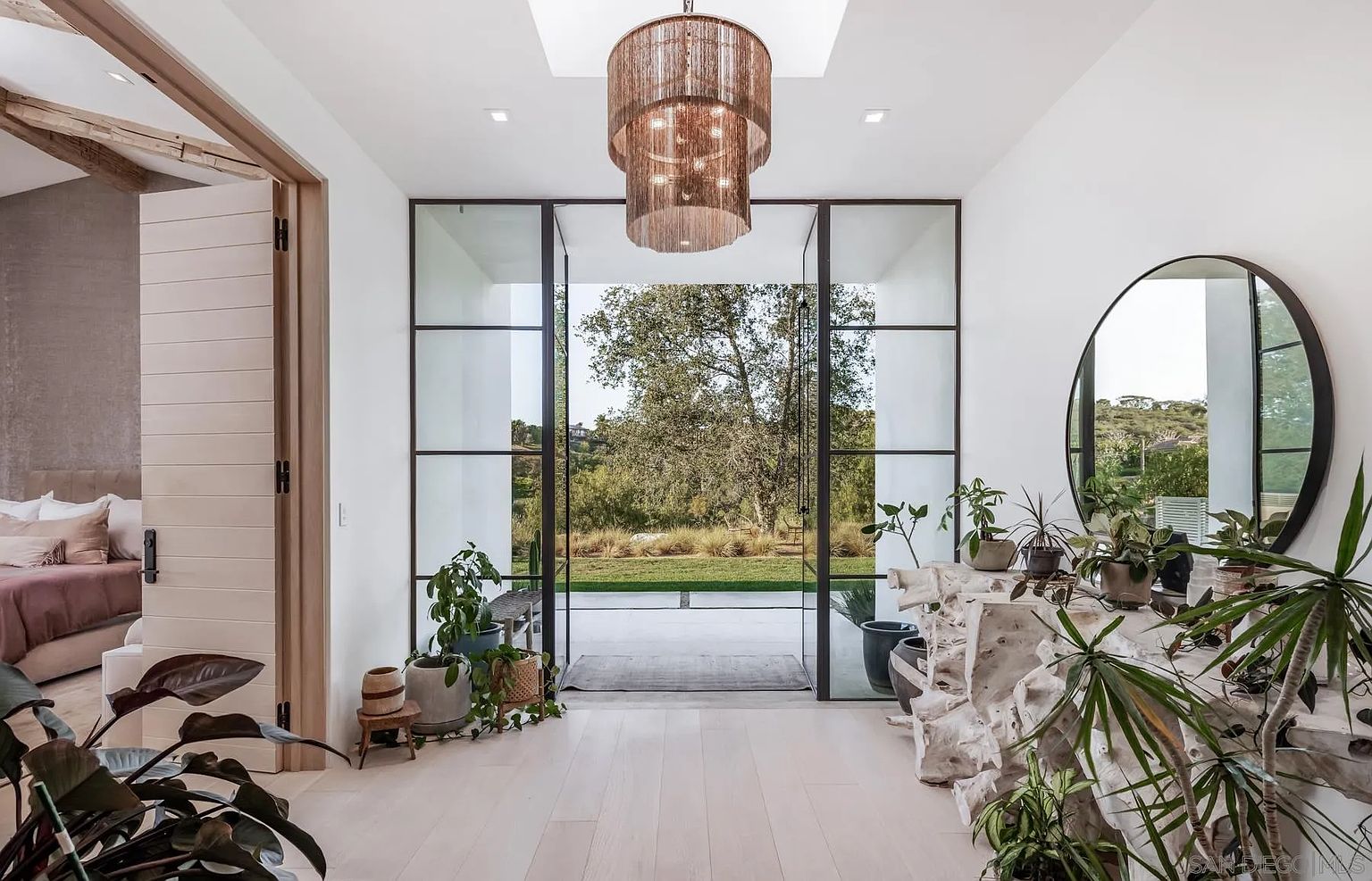 This bright and airy entryway features a striking chandelier hanging from a skylight, casting a warm glow over the light wood flooring. A large, circular mirror hangs above a unique, organic-shaped light wood console table adorned with various potted plants, creating a natural and inviting atmosphere. The space opens up through large glass doors to a scenic view of the lush green landscape outside, seamlessly blending indoor and outdoor living.