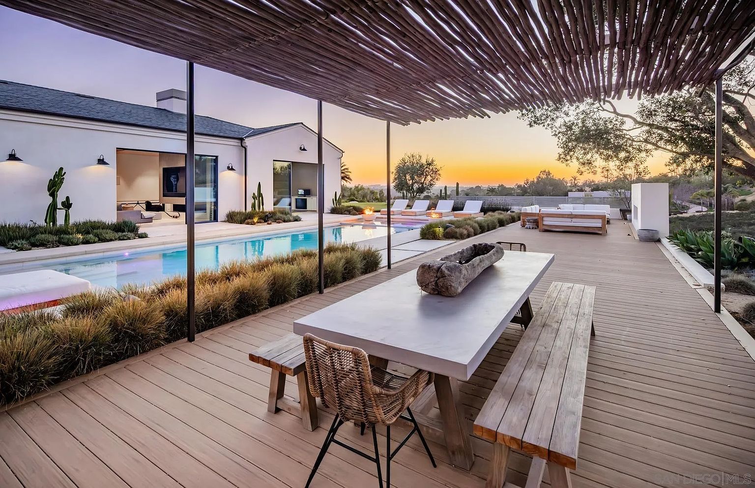 This expansive outdoor living space features a large wooden deck covered by a rustic, slatted wood pergola. The area includes a long dining table with benches and a woven chair, overlooking a sleek swimming pool and a sunset horizon. The modern home architecture is complemented by minimalist landscaping and a comfortable lounge area with white seating near the pool's edge.