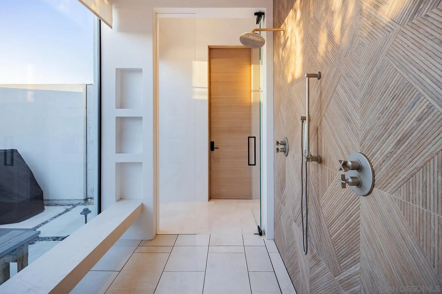 This modern bathroom features a walk-in shower with a striking geometric patterned tile wall and a gold-toned shower head. A built-in bench sits adjacent to a large glass panel that looks out onto a private outdoor space, while a light wood door provides access to the rest of the home. The space is characterized by its clean lines, neutral color palette, and seamless transition between indoor and outdoor elements.