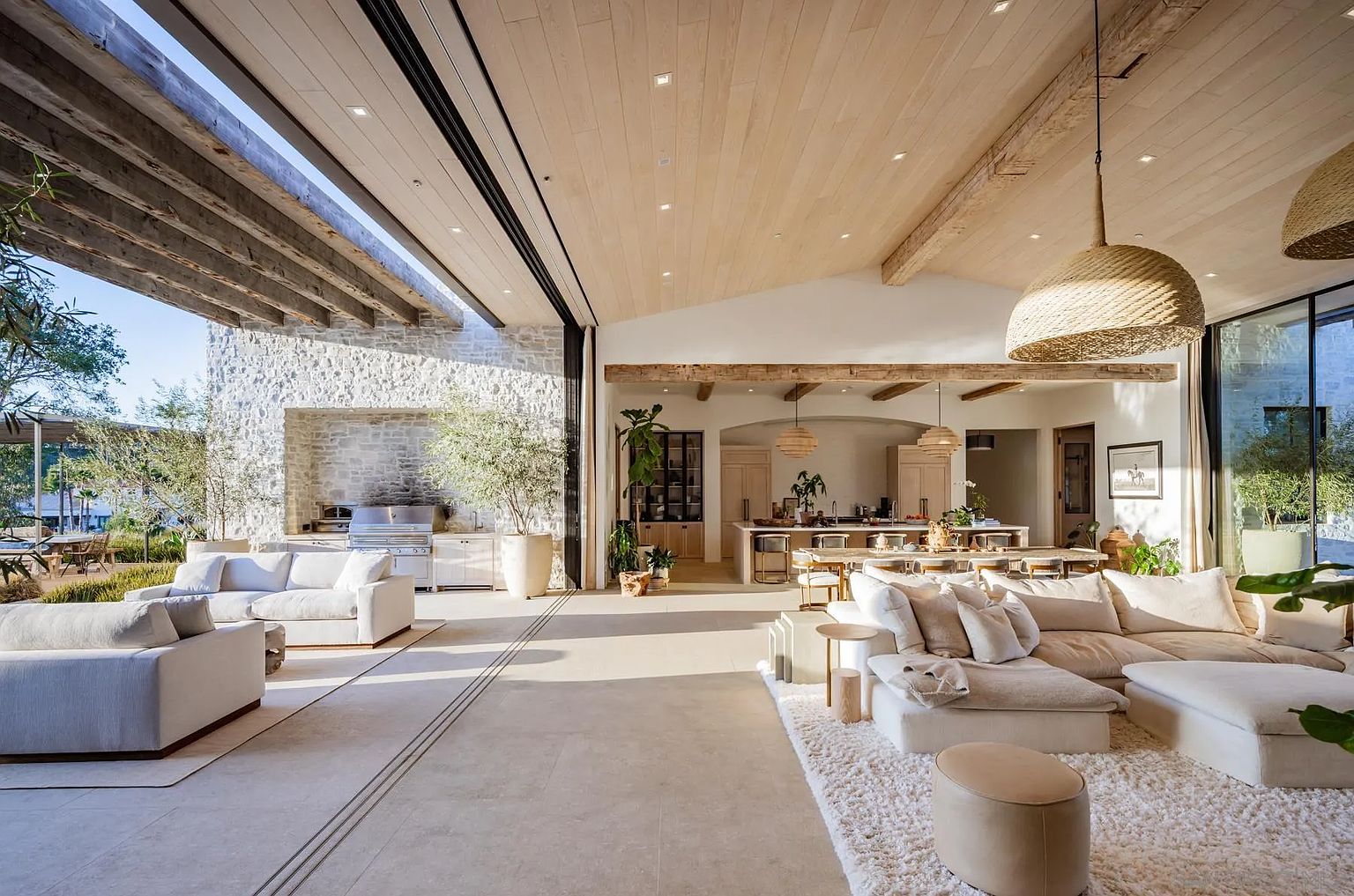 This expansive open-concept living space seamlessly blends indoor and outdoor living through massive sliding glass walls that open to a stone-clad patio. The interior features light wood ceilings with exposed beams, a neutral color palette, and a large, plush sectional arranged on a textured area rug. In the background, a bright kitchen and dining area complete the airy, sophisticated design, creating a cohesive and luxurious atmosphere.