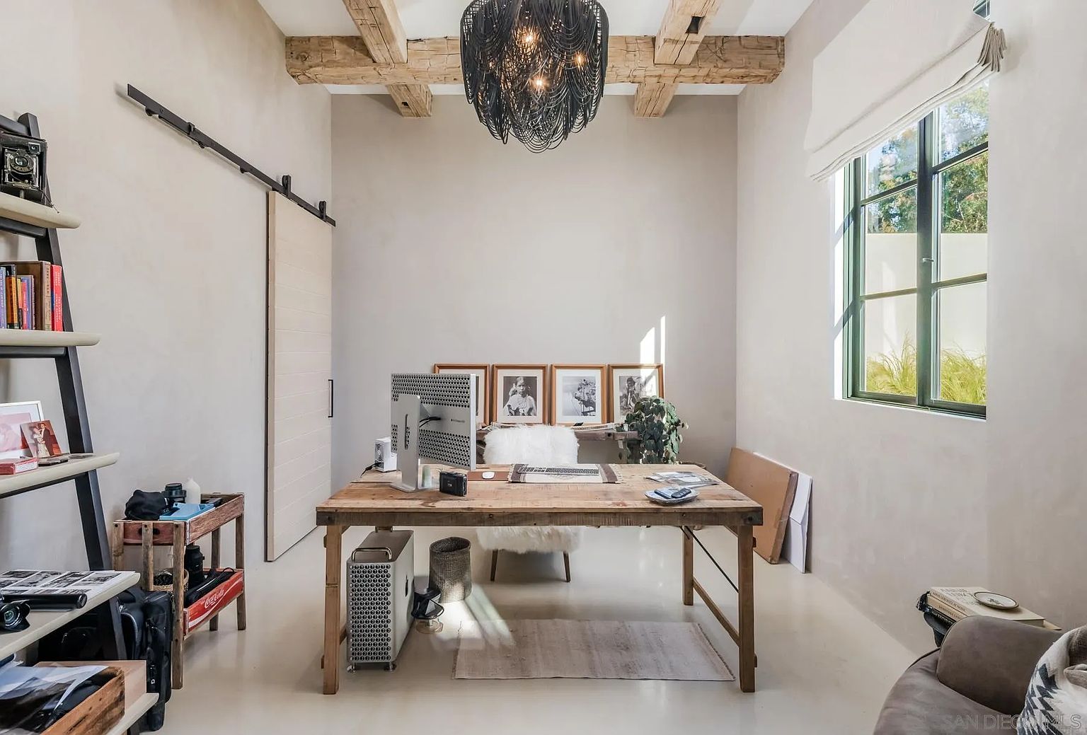 This home office features a rustic aesthetic with exposed wooden ceiling beams and a large, natural-wood desk centered in the room. A striking black chandelier hangs above the workspace, while a sliding barn door and a tall window provide character and natural light. The room is furnished with a white faux-fur chair and a shelving unit, creating a professional yet cozy atmosphere.