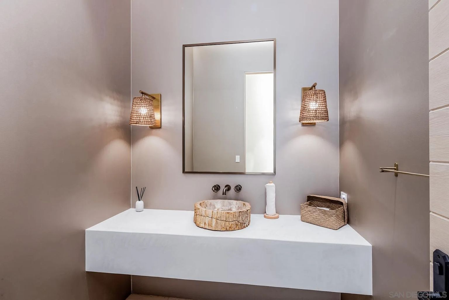 This modern powder room features a floating white vanity topped with a unique, natural-edge stone vessel sink. The space is accented by two woven-texture wall sconces flanking a rectangular mirror, creating a warm and organic aesthetic against the neutral-toned walls. A small woven basket and a roll of paper towels sit on the vanity, emphasizing the room's clean and minimalist design.