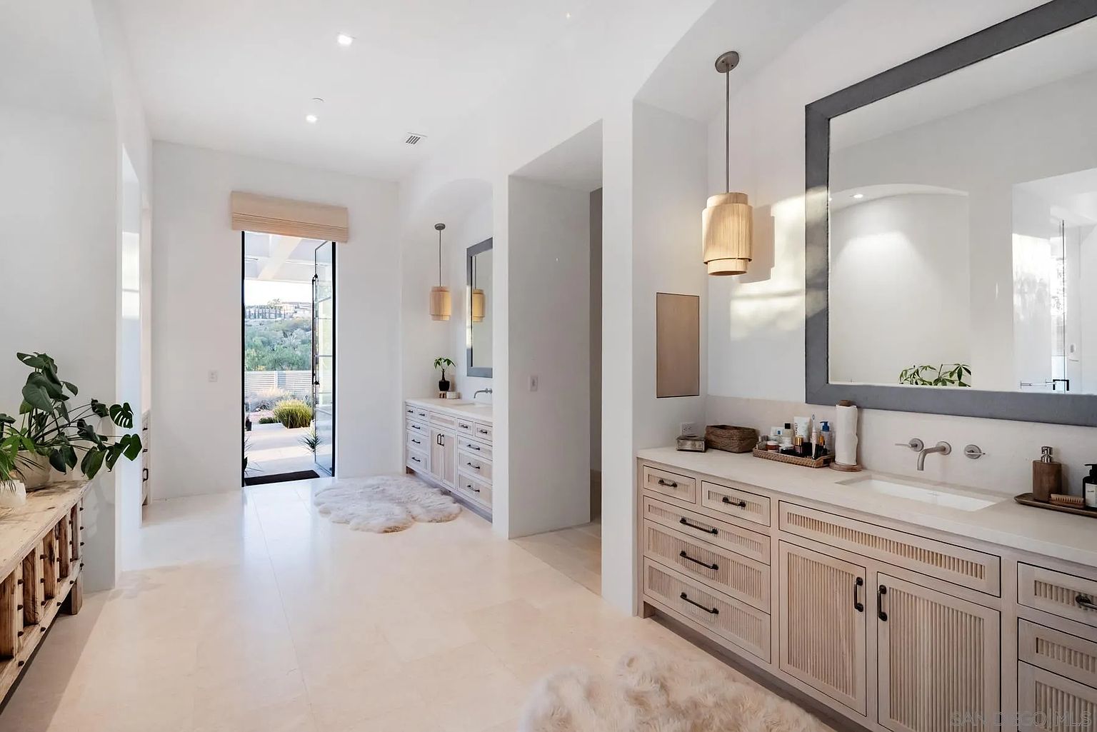 This spacious, light-filled primary bathroom features dual light-wood vanities with textured drawer fronts and neutral stone countertops. The room is accented by warm pendant lighting, large mirrors, and soft, fluffy rugs on the light-toned tile floor. A doorway at the end of the hall leads directly to an outdoor patio, creating a seamless transition between the indoor sanctuary and the natural landscape.