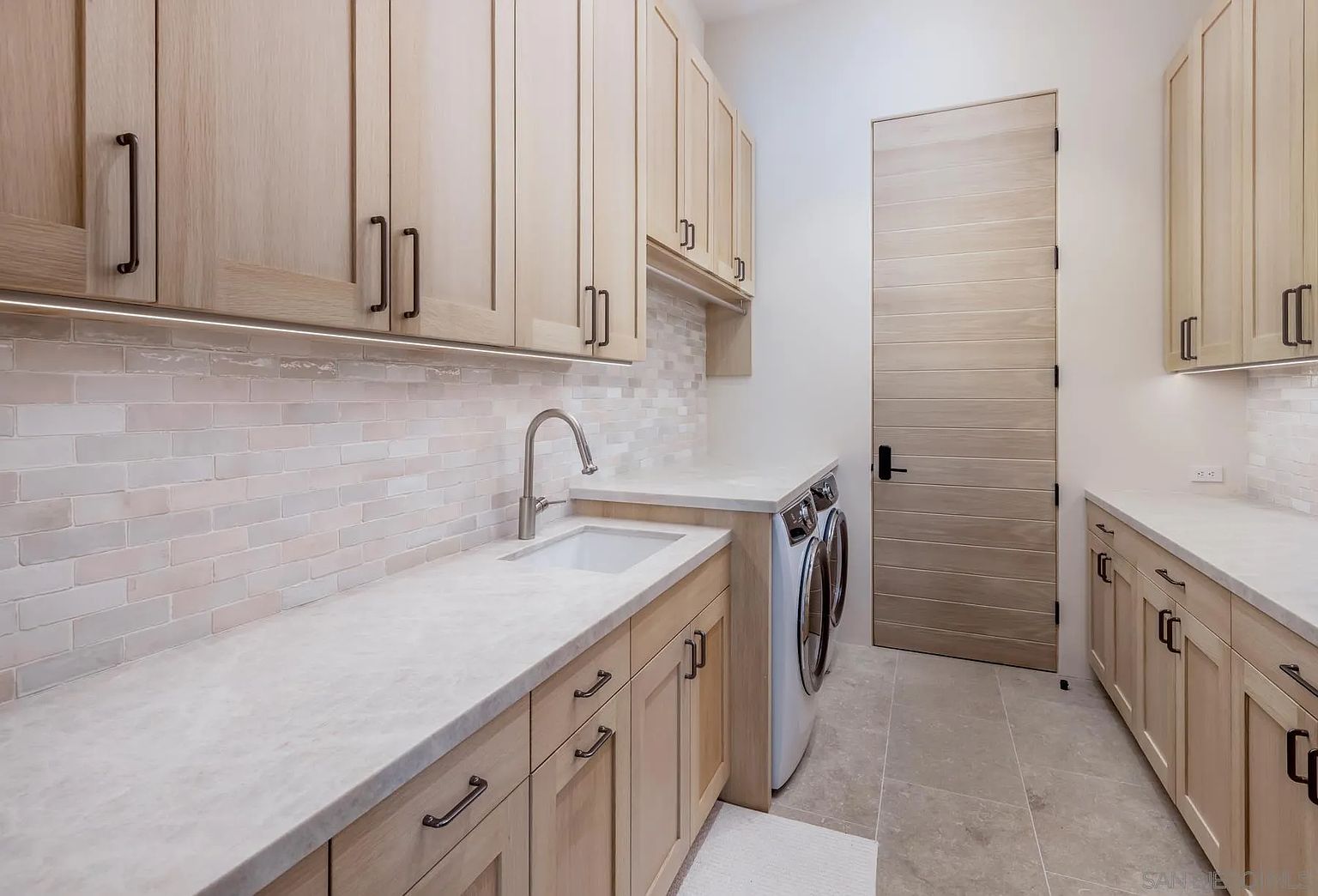 This modern laundry room features light wood cabinetry, a stone countertop with an integrated sink, and a tiled backsplash. The space is equipped with a side-by-side washer and dryer set and is finished with neutral-toned floor tiles and a matching wood-paneled door. The clean, organized layout provides a functional and aesthetically pleasing workspace.