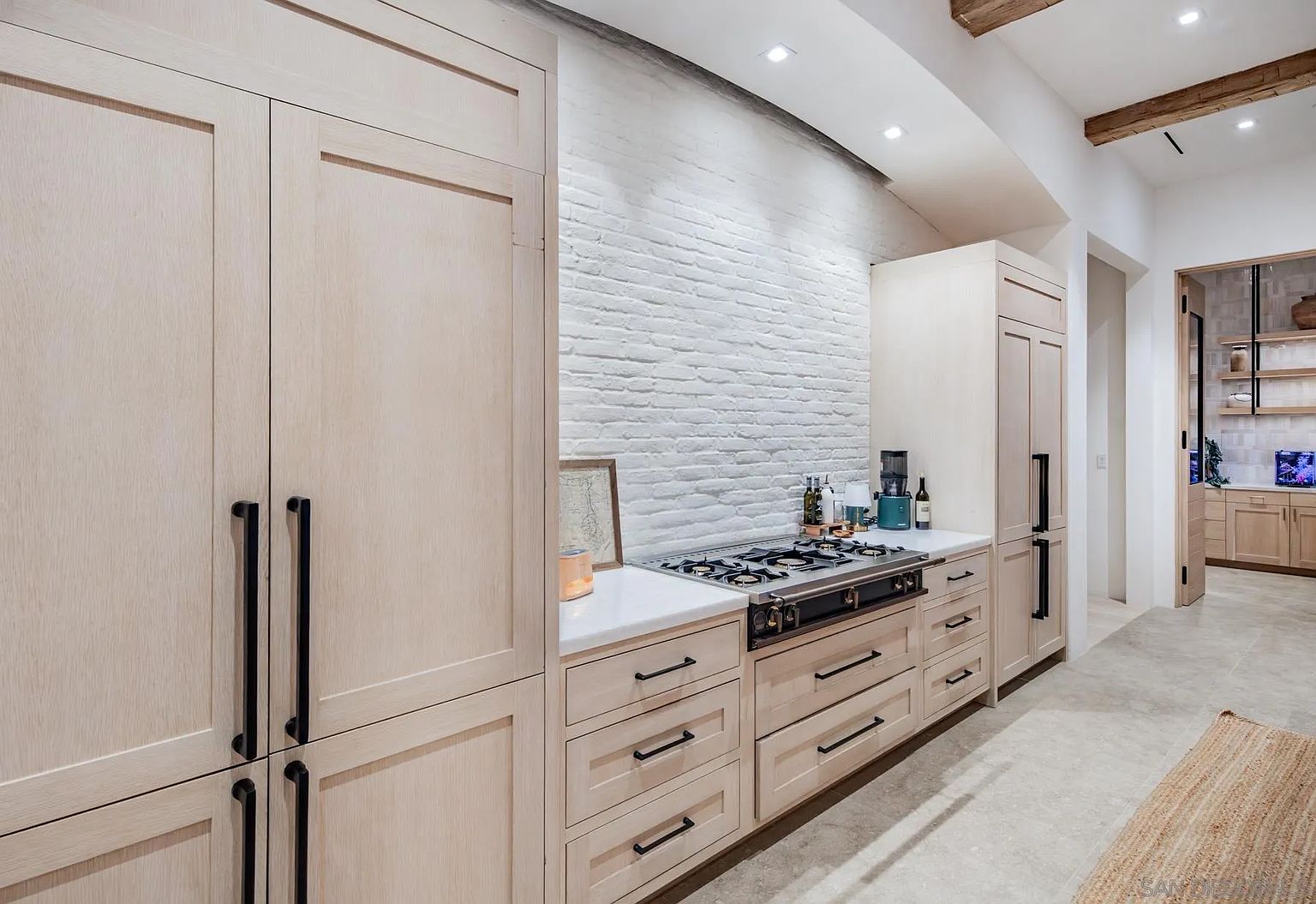 This modern kitchen features light wood cabinetry with matte black hardware and a prominent white brick backsplash. A professional-style gas range is integrated into the cabinetry, complemented by light-colored stone countertops and a neutral tile floor. The perspective captures the clean, minimalist aesthetic of the cooking area, with a glimpse into an adjacent room featuring open shelving.