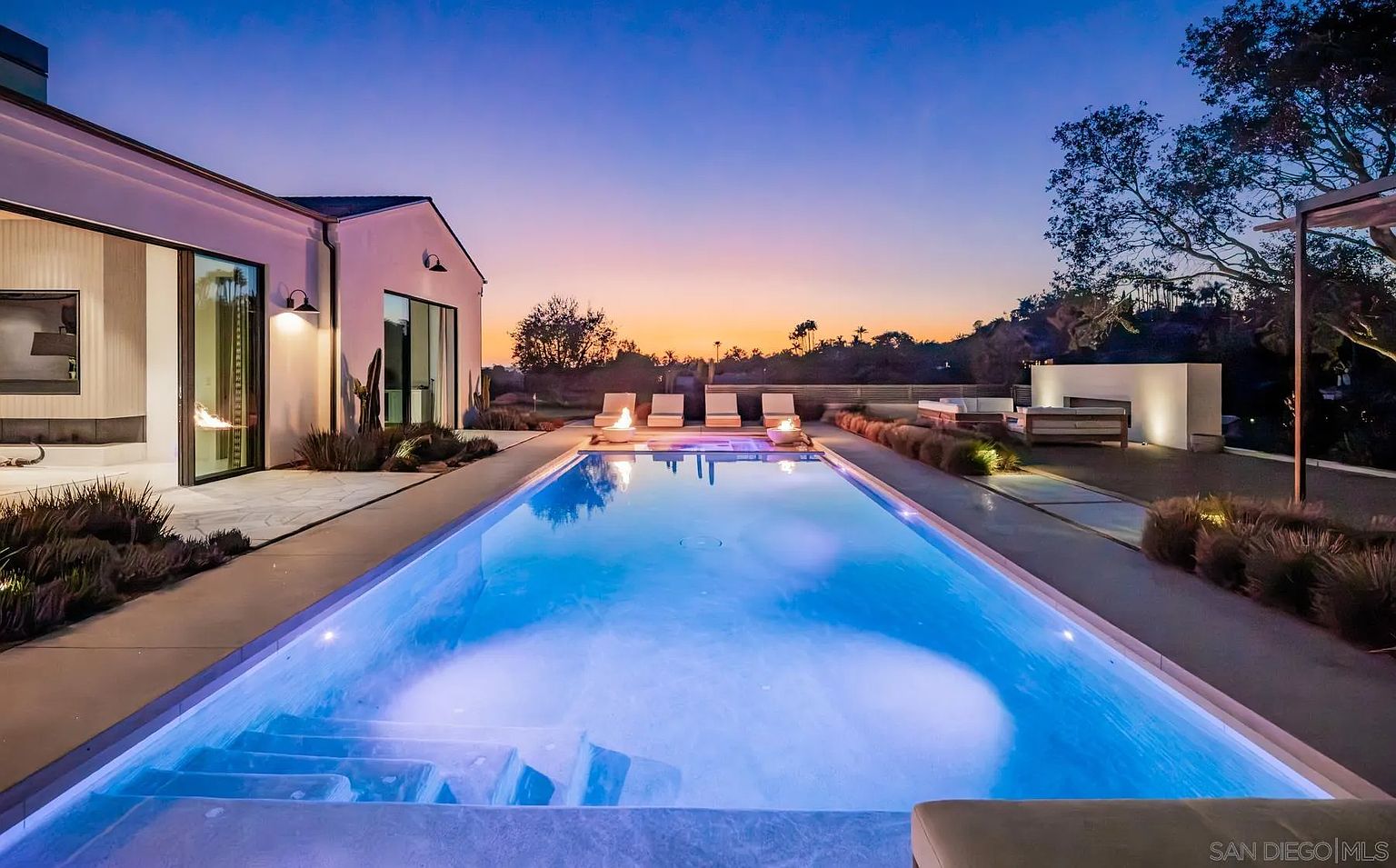 This stunning backyard features a long, rectangular swimming pool as the central focal point, illuminated by soft blue underwater lighting at dusk. The surrounding patio is paved with large, neutral-toned tiles and includes a row of lounge chairs and a separate seating area with a modern outdoor fireplace. The scene is framed by a contemporary home with large glass sliding doors, creating a seamless transition between indoor and outdoor living spaces.