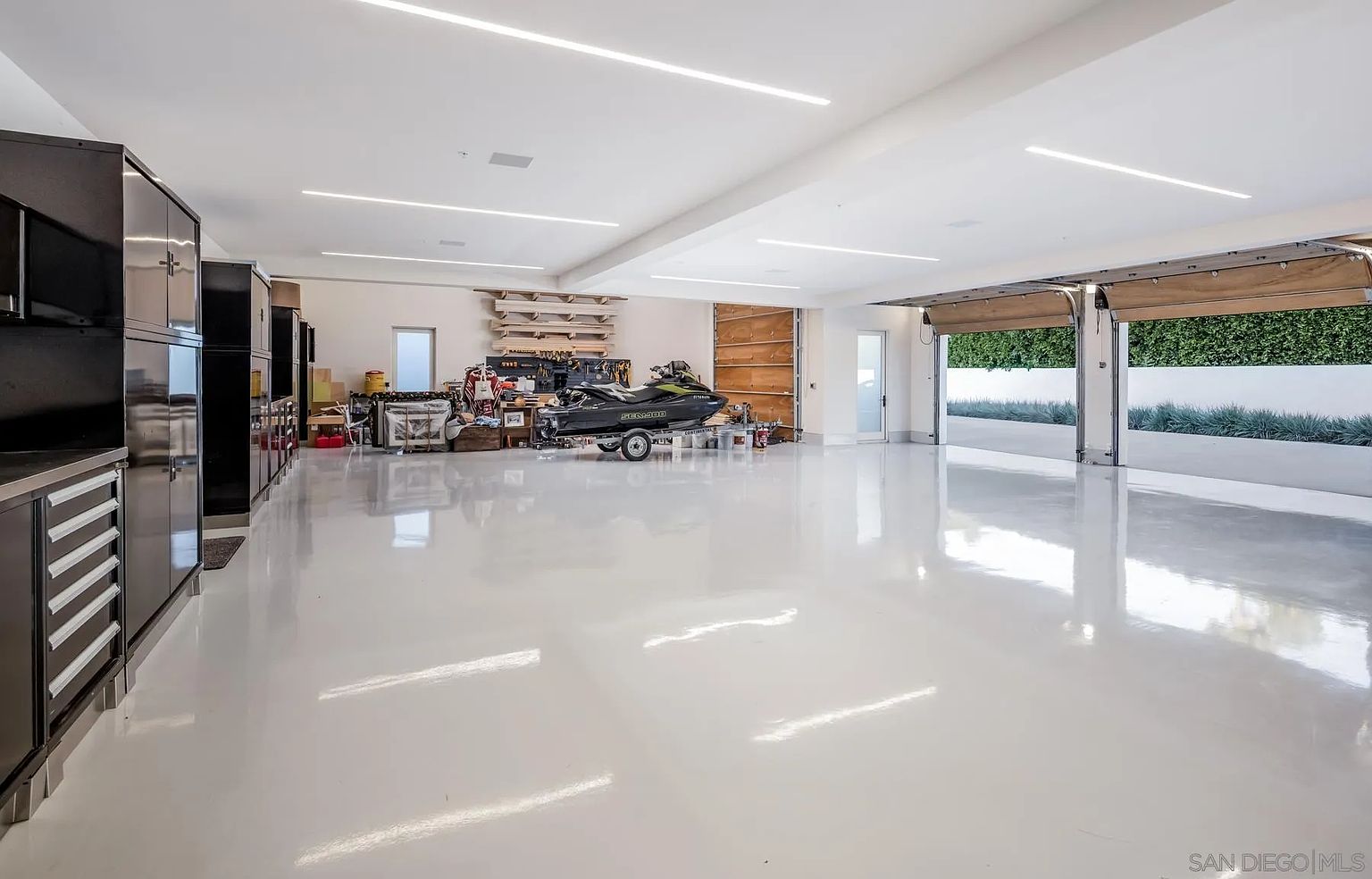 This expansive, modern garage features a pristine, high-gloss white epoxy floor and bright, linear recessed lighting that creates a clean, professional aesthetic. The space is equipped with extensive dark cabinetry for storage, a dedicated workbench area, and a jet ski on a trailer, highlighting its versatility for both vehicle storage and hobbyist projects. Large, open garage doors reveal a view of a manicured hedge and outdoor landscaping, seamlessly connecting the interior workspace to the exterior environment.