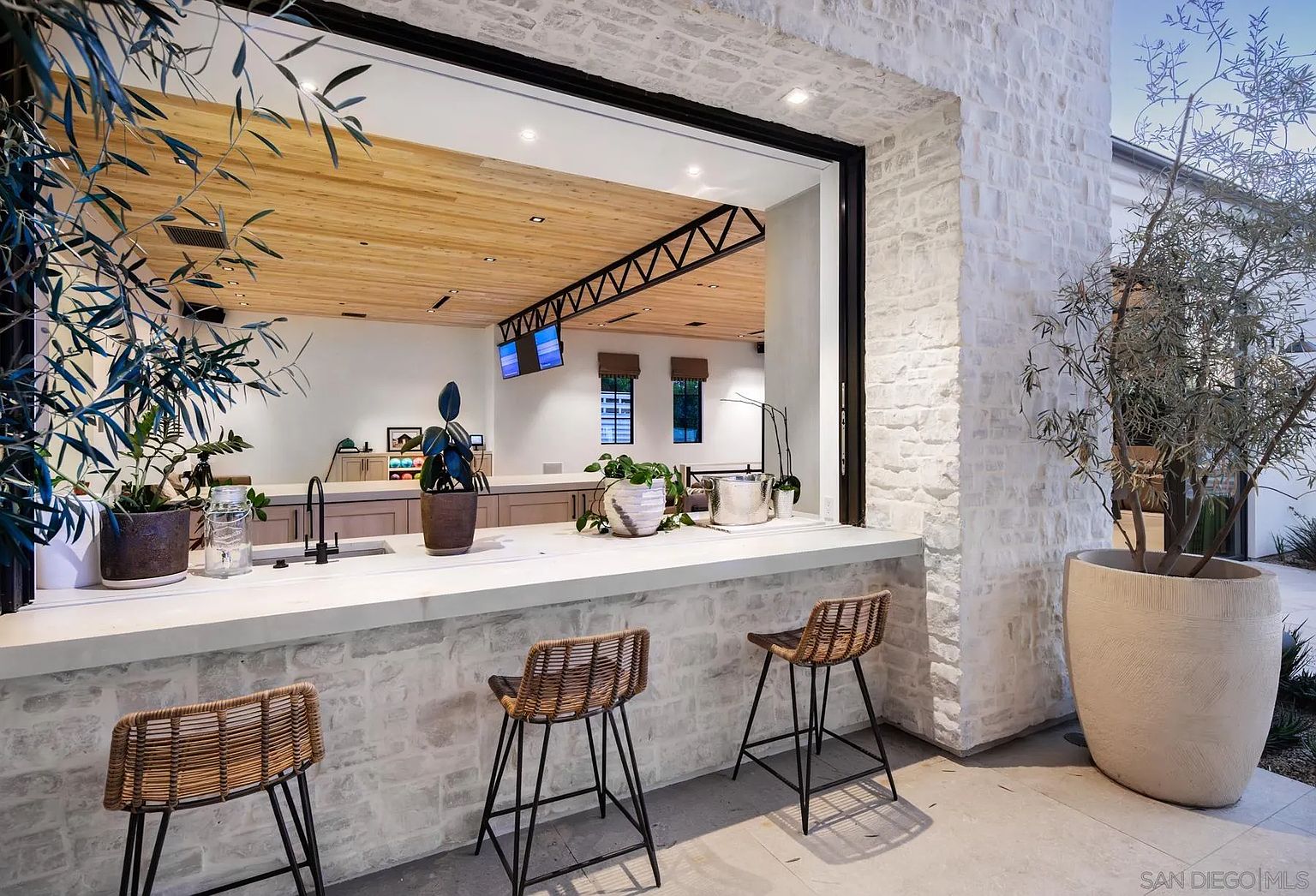 This outdoor patio space features a seamless transition to the interior through a large, open window pass-through with a stone-topped bar counter. Three woven bar stools are positioned along the exterior side, while a large potted olive tree adds a touch of greenery to the stone-clad wall. The view captures a sophisticated indoor-outdoor living area, highlighting the open-concept design and modern architectural details.