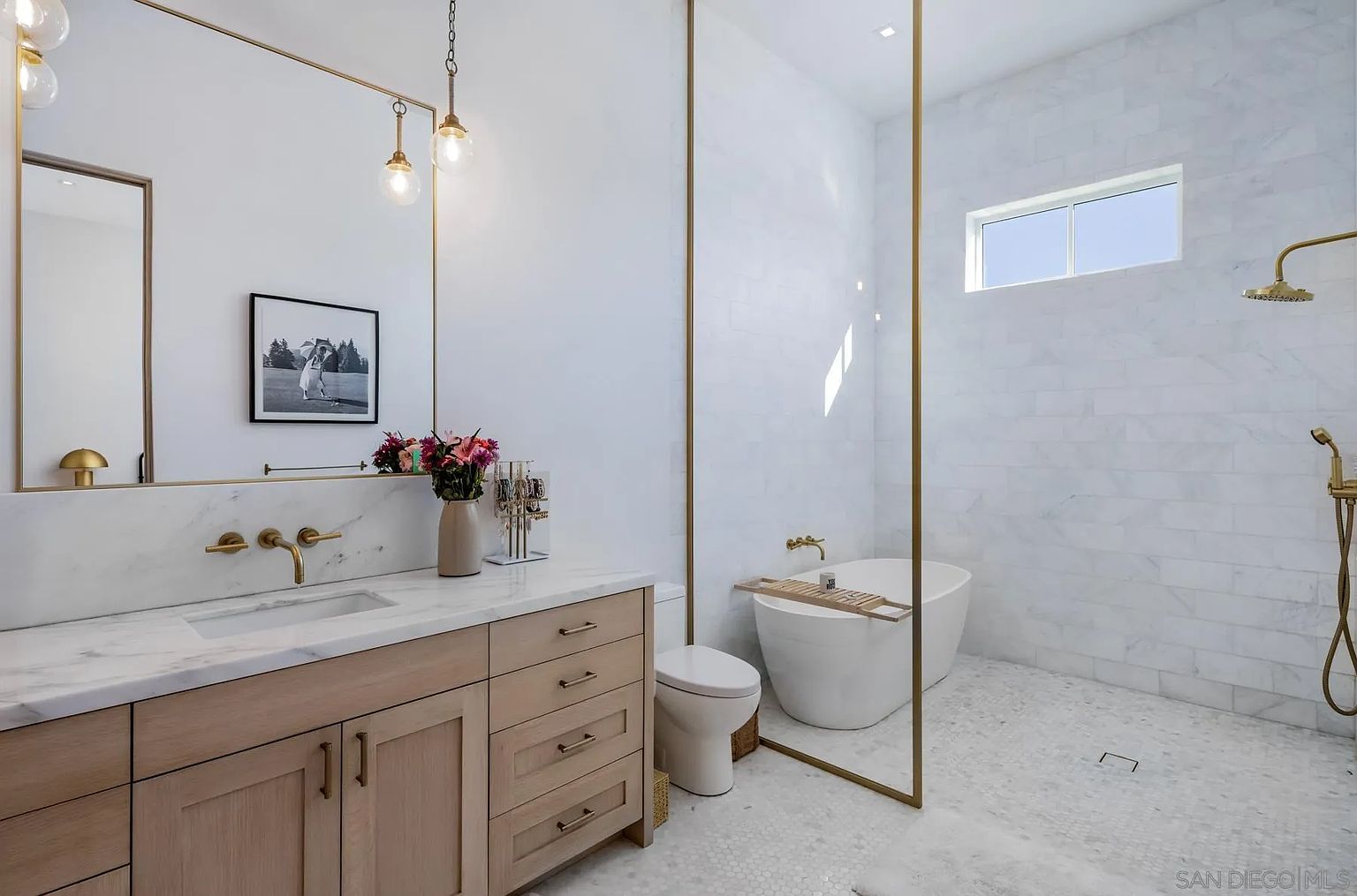 This modern primary bathroom features a light wood vanity with a stone countertop and a large, gold-framed mirror. A freestanding soaking tub is positioned within a wet room area, enclosed by a tall gold frame and glass partition, complemented by a wall-mounted shower fixture. The space is finished with light-toned wall and floor tiles, creating a bright, clean, and luxurious atmosphere.