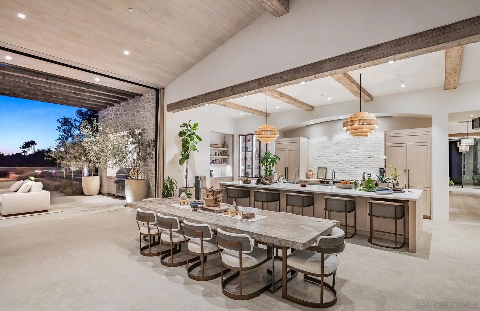 This expansive, open-concept kitchen and dining area features a seamless transition to an outdoor living space through a massive wall of glass. The room is anchored by a long dining table and a large kitchen island with bar seating, both illuminated by textured pendant lights and exposed wooden ceiling beams. The aesthetic is sophisticated and airy, characterized by light-toned stone flooring, neutral cabinetry, and a textured white brick wall behind the kitchen workspace.