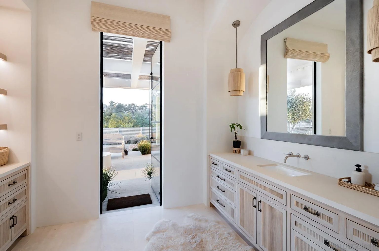 This bright and airy bathroom features a long vanity with light wood cabinetry, a white countertop, and a large mirror framed in a textured gray finish. A tall glass door provides direct access to an outdoor patio, blending indoor and outdoor living spaces. The room is accented with a soft rug, a pendant light, and a small potted plant, creating a serene and modern atmosphere.