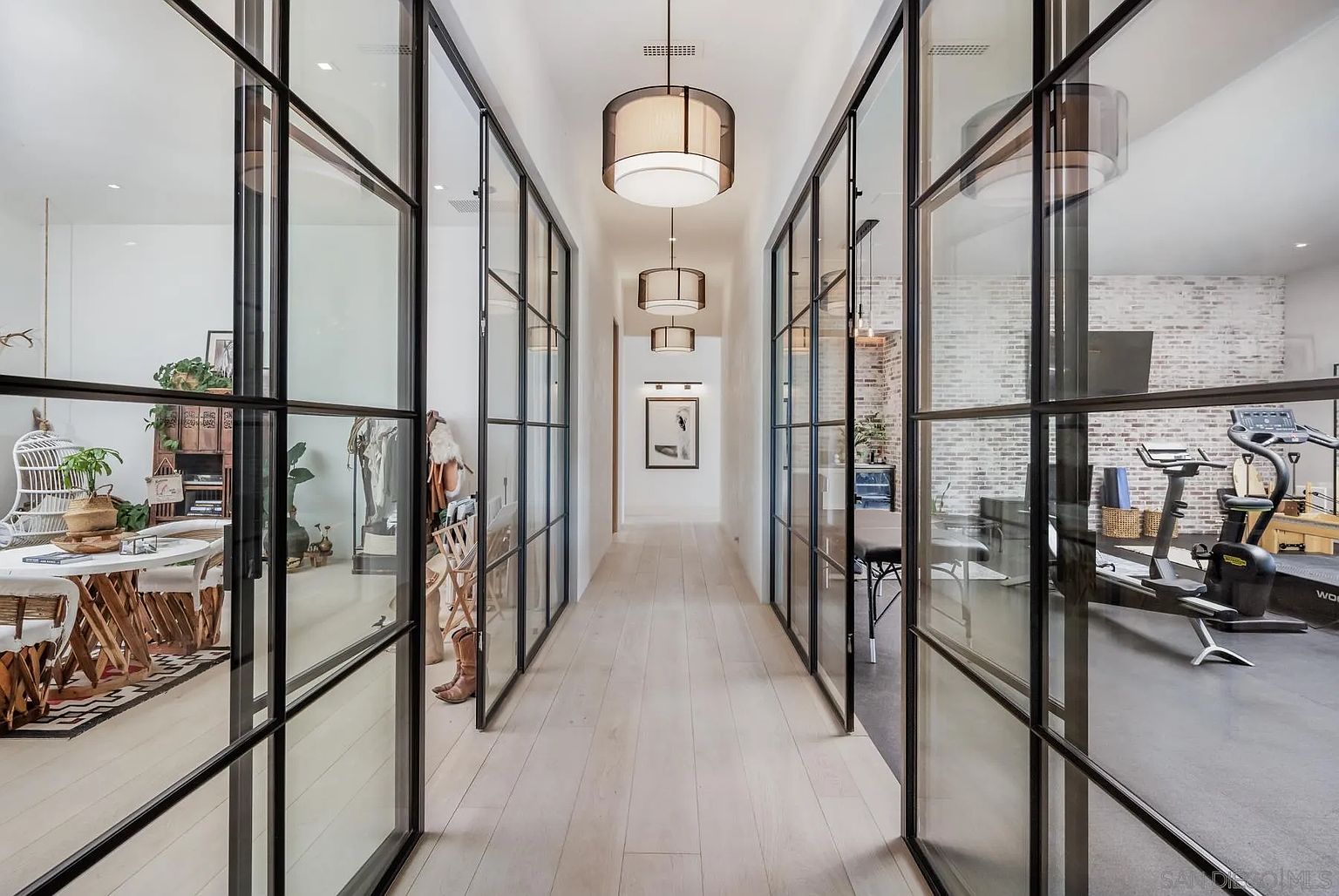 This perspective captures a long, symmetrical hallway featuring light wood flooring and a series of three modern, drum-style pendant lights. The space is defined by large, black-framed glass partitions that provide a clear view into adjacent rooms, including a dining area on the left and a home gym on the right. The clean lines and minimalist aesthetic create a sophisticated, open-concept transition between living spaces.