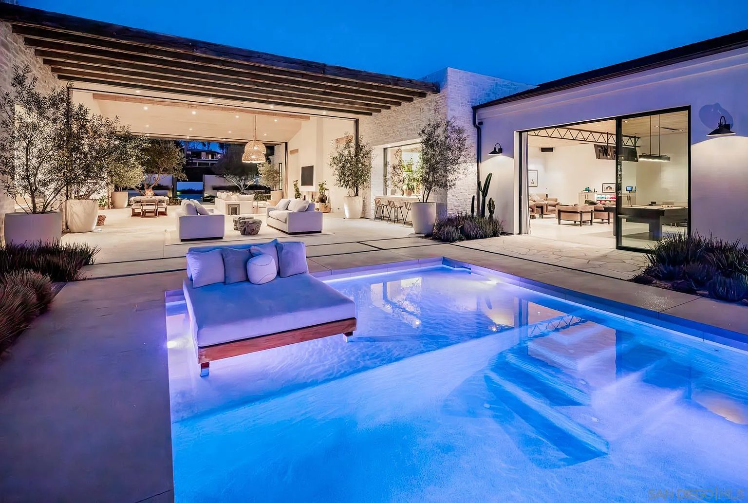 This stunning outdoor living space features a modern swimming pool with a built-in lounge platform, set against a backdrop of a contemporary home with expansive open-air living areas. The architecture emphasizes seamless indoor-outdoor flow, with large sliding glass doors connecting the interior rooms to a spacious patio area illuminated by warm ambient lighting. The scene is captured at twilight, highlighting the vibrant blue glow of the pool and the sophisticated, resort-like atmosphere of the property.