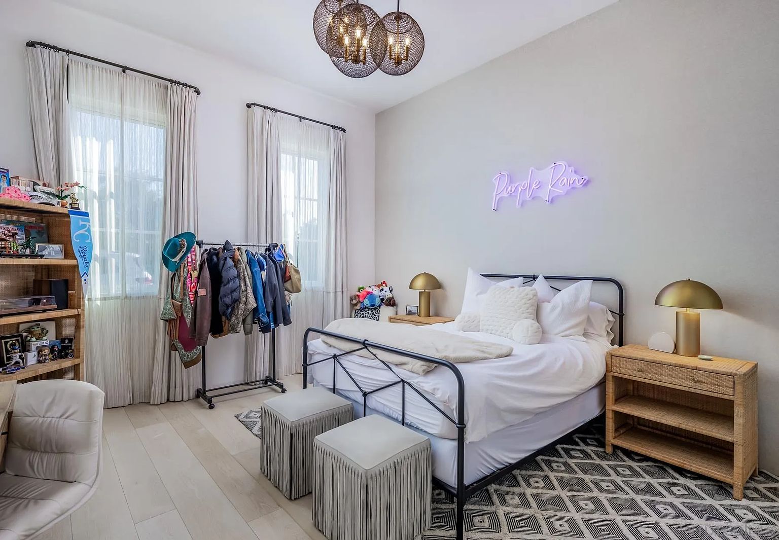 This bedroom features a black metal bed frame centered against a light-toned wall, accented by a neon 'Purple Rain' sign. The room includes a woven nightstand, a clothing rack, and a tall wooden shelving unit, all set upon a geometric patterned rug. The space is illuminated by a unique multi-sphere pendant light and two gold-toned table lamps, creating a cozy and eclectic atmosphere.