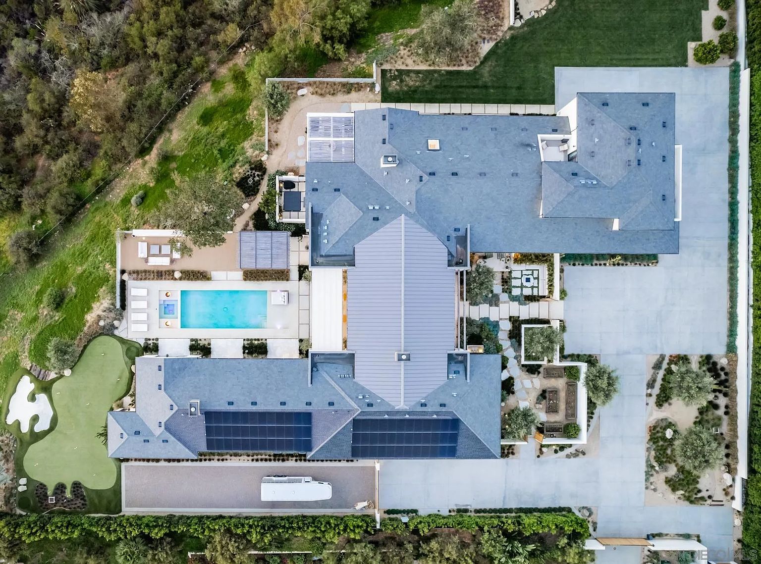 This high-altitude aerial view captures a sprawling luxury estate, showcasing its complex roofline, solar panel arrays, and expansive outdoor living spaces. The property features a rectangular swimming pool, a putting green, and a large paved driveway, all set against a backdrop of lush hillside vegetation. The layout emphasizes a seamless integration of indoor and outdoor living areas, highlighting the property's significant footprint and private grounds.