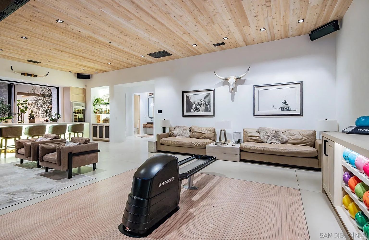 This unique interior space features a private bowling alley as its centerpiece, complete with a Brunswick ball return and a rack of colorful bowling balls. The room is designed for entertainment, incorporating a lounge area with tan leather sofas, a bar with high-top seating, and a light wood-paneled ceiling. Western-inspired decor, including two large animal skulls mounted on the walls, adds a distinct character to this spacious, open-concept recreational room.