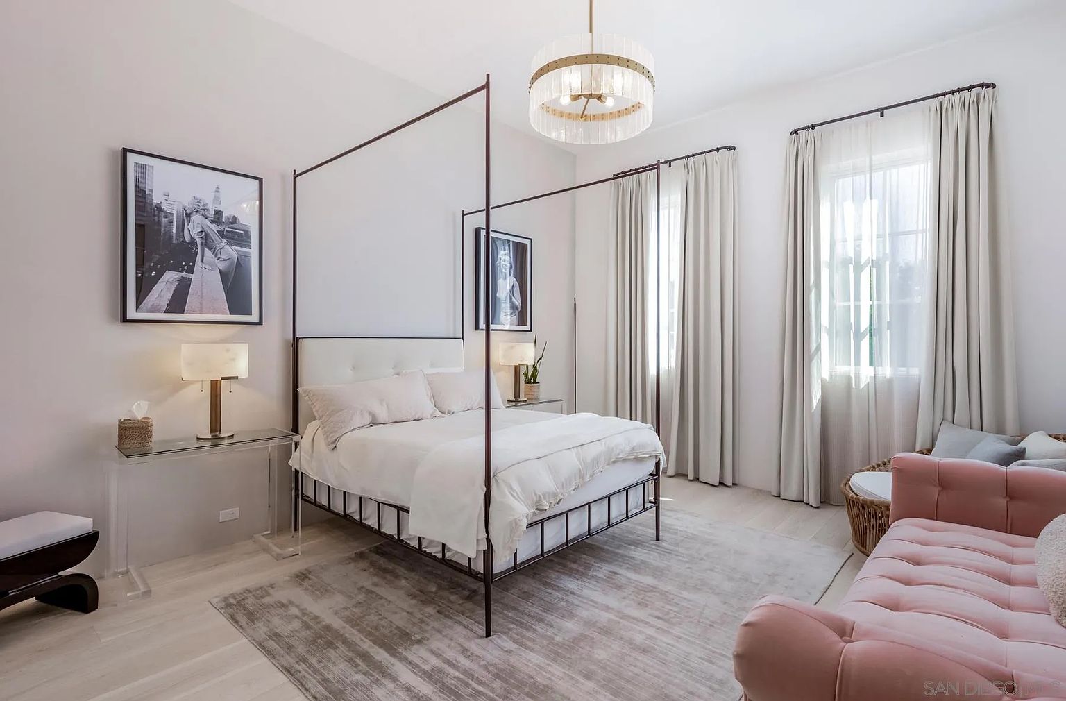 This elegant primary bedroom features a dark metal canopy bed as the central focal point, complemented by a soft, neutral color palette and light wood flooring. A plush pink tufted bench sits at the foot of the bed, while large windows draped in floor-to-ceiling curtains allow ample natural light to fill the space. The room is accented by a sophisticated crystal chandelier and framed black-and-white photography, creating a serene and luxurious atmosphere.