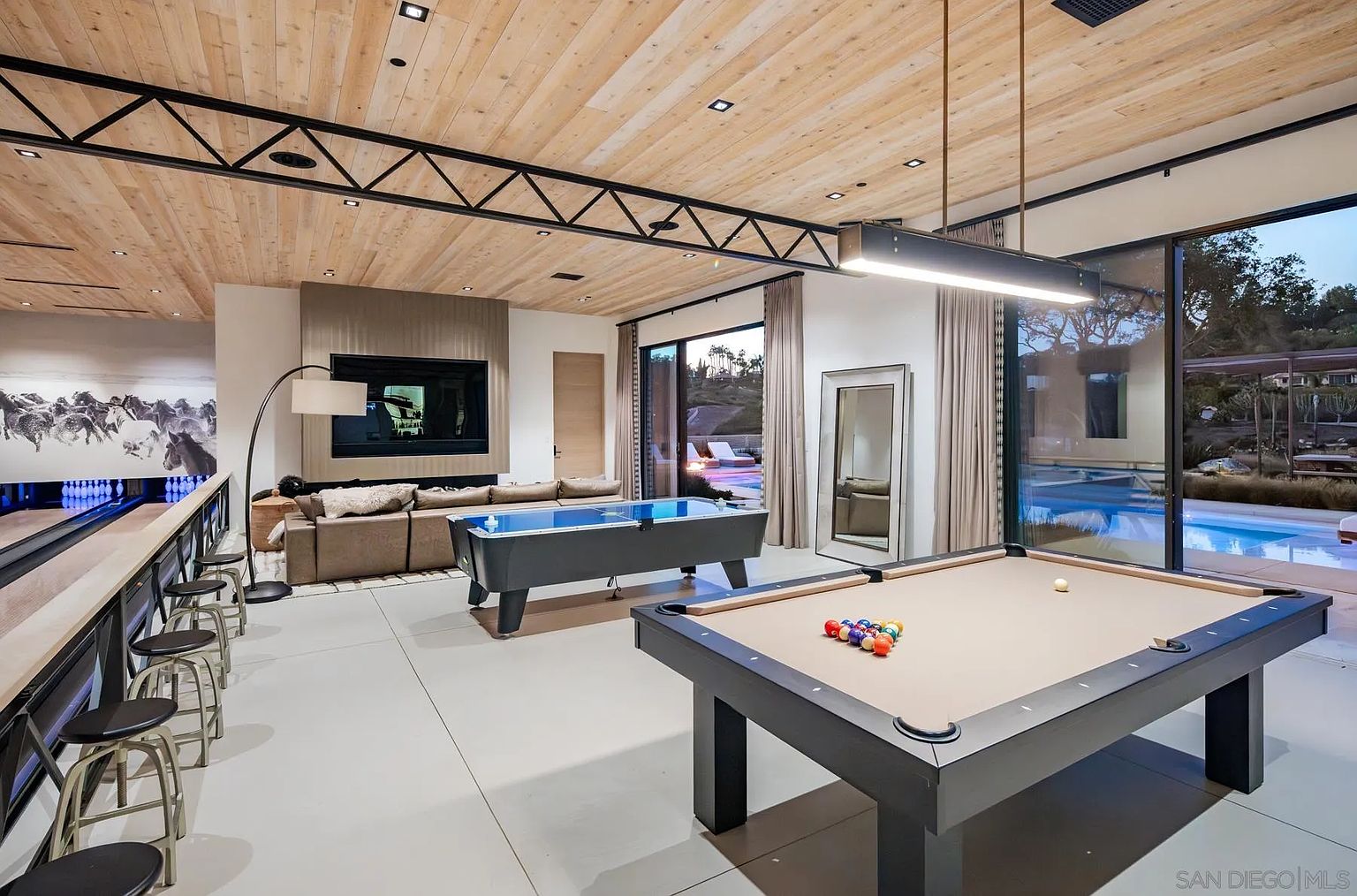 This expansive game room features a modern aesthetic with a light wood-paneled ceiling, a prominent black metal truss, and a polished concrete floor. The space is equipped with a pool table, an air hockey table, and a bowling lane, alongside a comfortable seating area with a sofa facing a wall-mounted television. Large glass sliding doors provide a seamless transition to an outdoor pool area, creating an open and airy atmosphere perfect for entertainment.