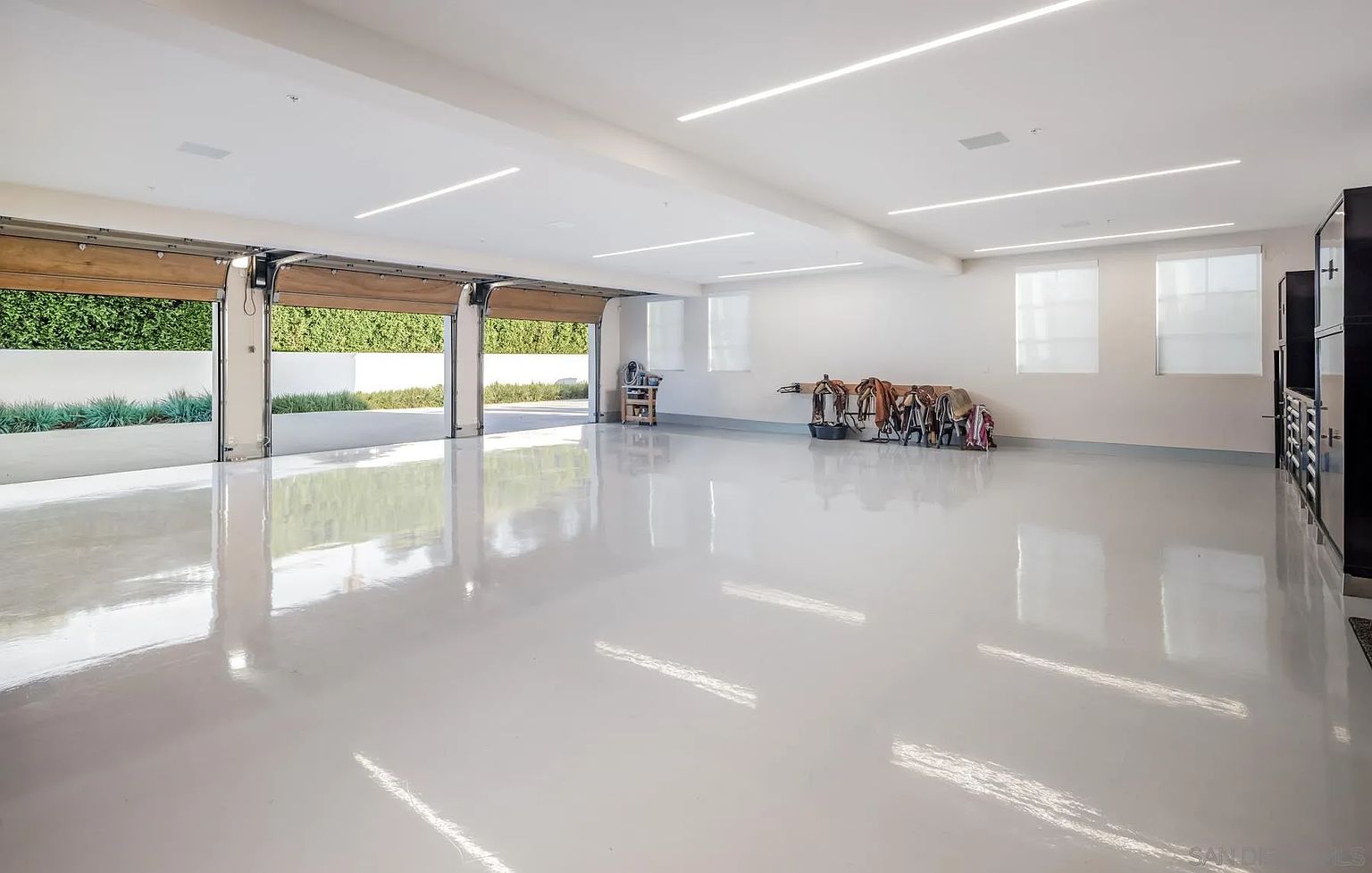 This expansive, pristine garage features a high-gloss, light-colored epoxy floor that reflects the overhead linear LED lighting. The space is open and airy, with three large, open garage door bays providing a seamless transition to the outdoor area. Along the back wall, a wooden rack holds several horse saddles, suggesting a functional space for equestrian storage or a multi-purpose workshop.