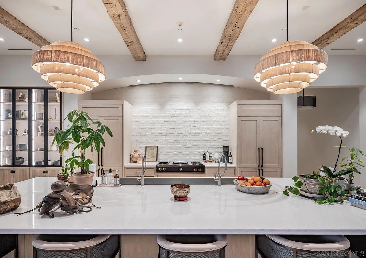 This sophisticated kitchen features a large central island with a stone countertop, serving as the focal point of the room. The space is defined by light wood cabinetry, a white brick backsplash behind the range, and exposed wooden ceiling beams that add a rustic touch. Two large, tiered pendant lights hang above the island, while a glass-front display cabinet on the left provides an elegant storage solution.