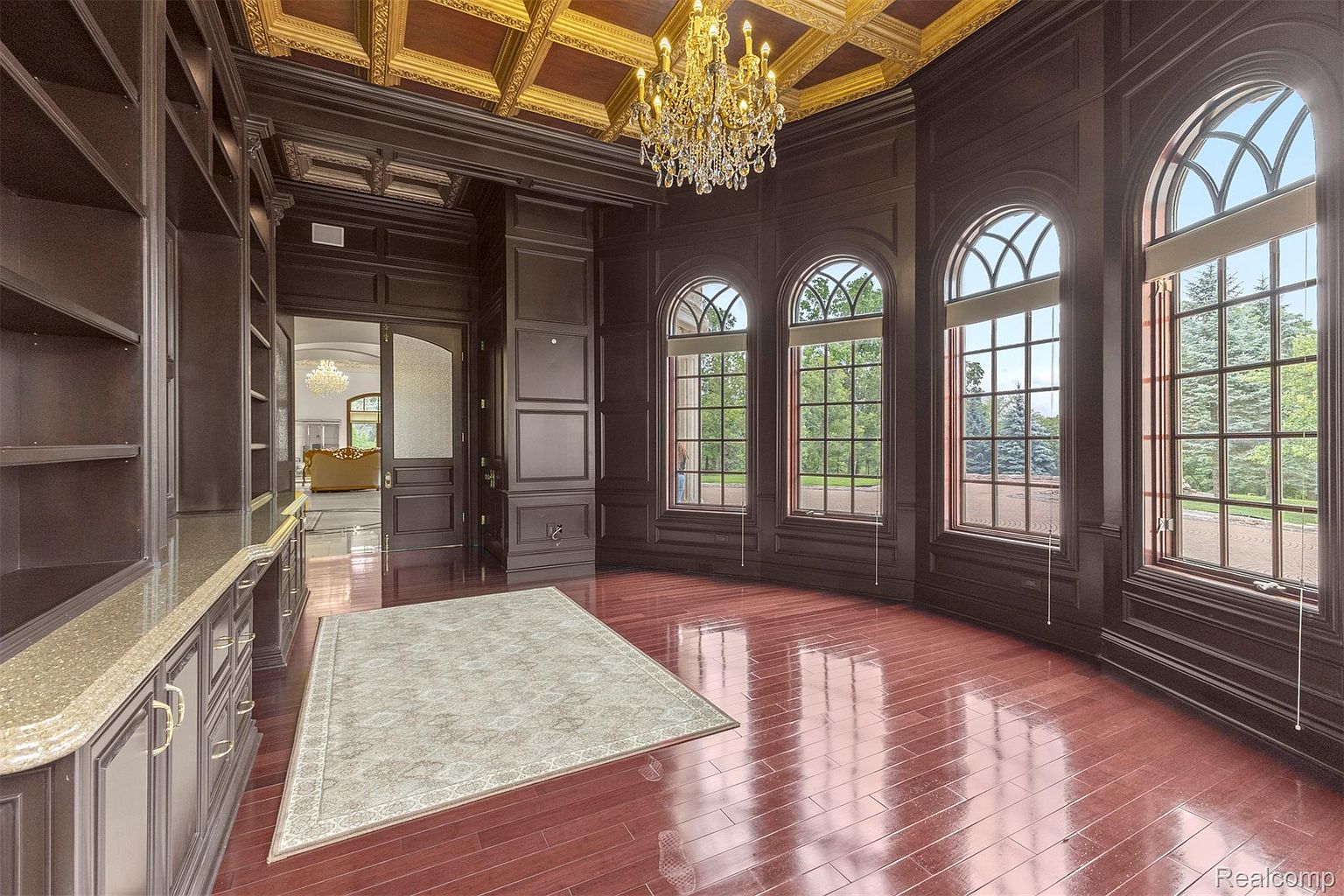 This is an interior shot of a luxurious office or study. The room features dark wood paneling, a coffered ceiling with gold accents, and a crystal chandelier. Large arched windows provide ample natural light, and a built-in bookcase offers storage. A decorative rug sits on the polished wood floor, adding a touch of elegance.