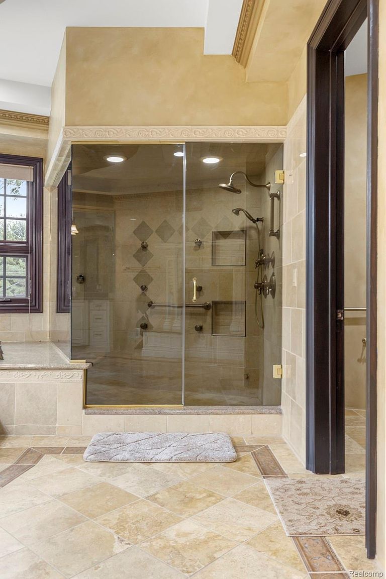 This is a luxurious primary bathroom featuring a glass-enclosed shower with bronze fixtures and tiled walls. The floor is tiled in a diamond pattern with a decorative border, and a plush bath mat sits in front of the shower. The overall impression is one of elegance and comfort.