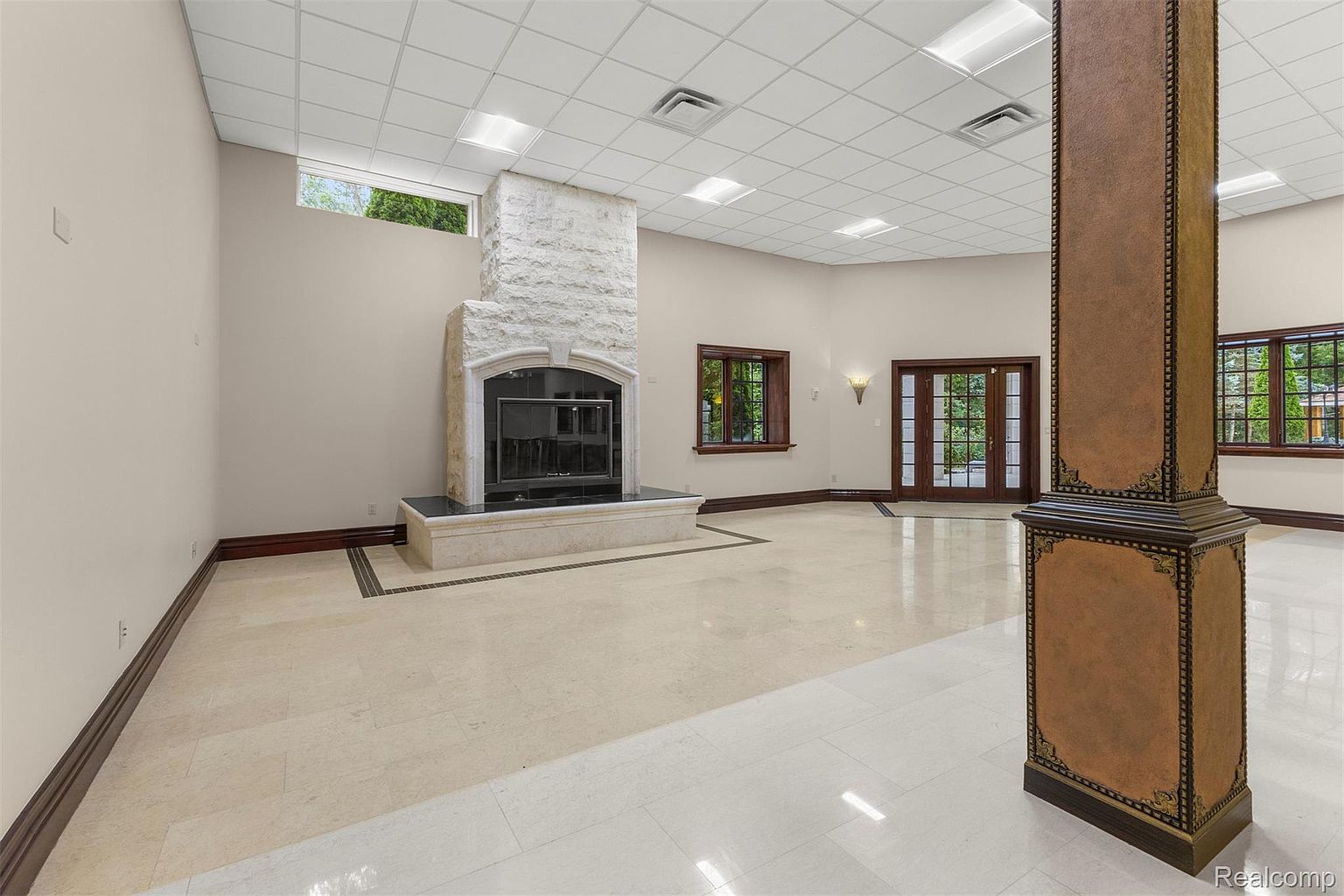This is a spacious living room featuring a stone fireplace as a focal point. The room has light-colored tile flooring, neutral walls, and dark wood trim. A decorative column stands prominently in the foreground, and the room is well-lit with recessed lighting and natural light from windows and glass doors.
