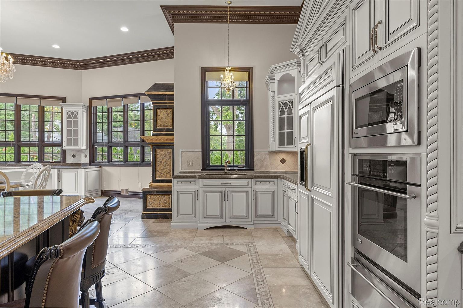 This is a luxurious kitchen featuring custom white cabinetry with detailed trim, stainless steel appliances including a built-in microwave and oven, and elegant countertops. A large window provides natural light, and the tile flooring adds to the sophisticated aesthetic. The kitchen exudes a sense of high-end craftsmanship and timeless design.