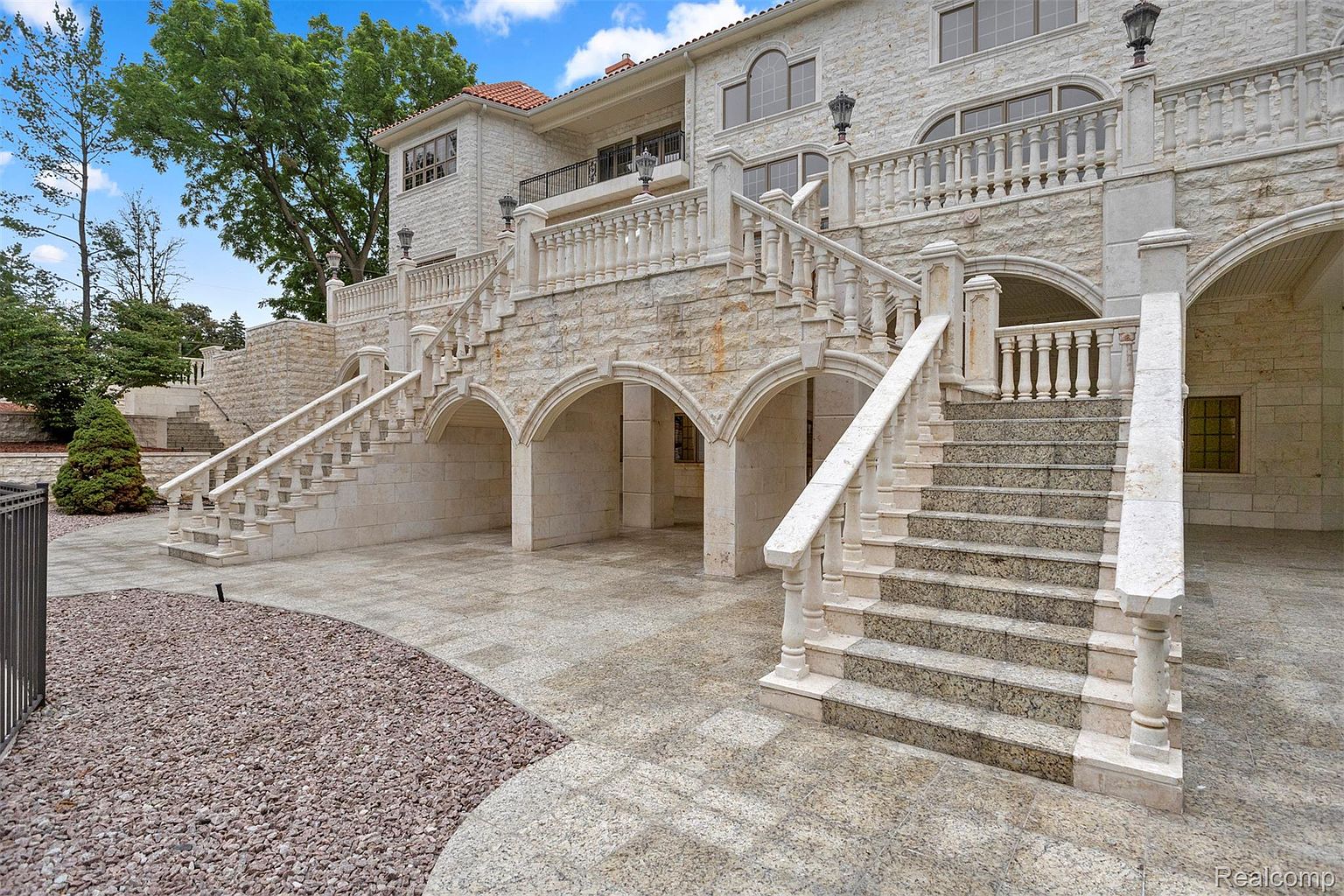 This image showcases the rear exterior of a grand, multi-story stone residence. The architecture features intricate stonework, arched entryways, and multiple staircases leading to upper levels. The landscaping includes a stone-paved patio area and gravel accents, creating a luxurious and inviting outdoor space.
