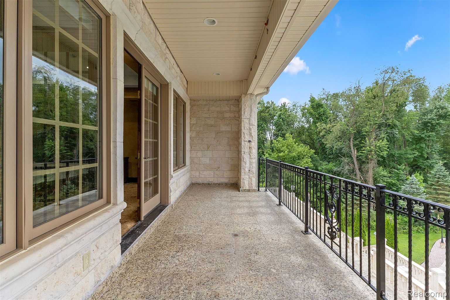 This image showcases a spacious balcony with a stone tile floor and a decorative black metal railing. The balcony is attached to a building with stone cladding and features large windows and a door leading inside. Lush greenery and trees are visible in the background, creating a serene and private atmosphere.