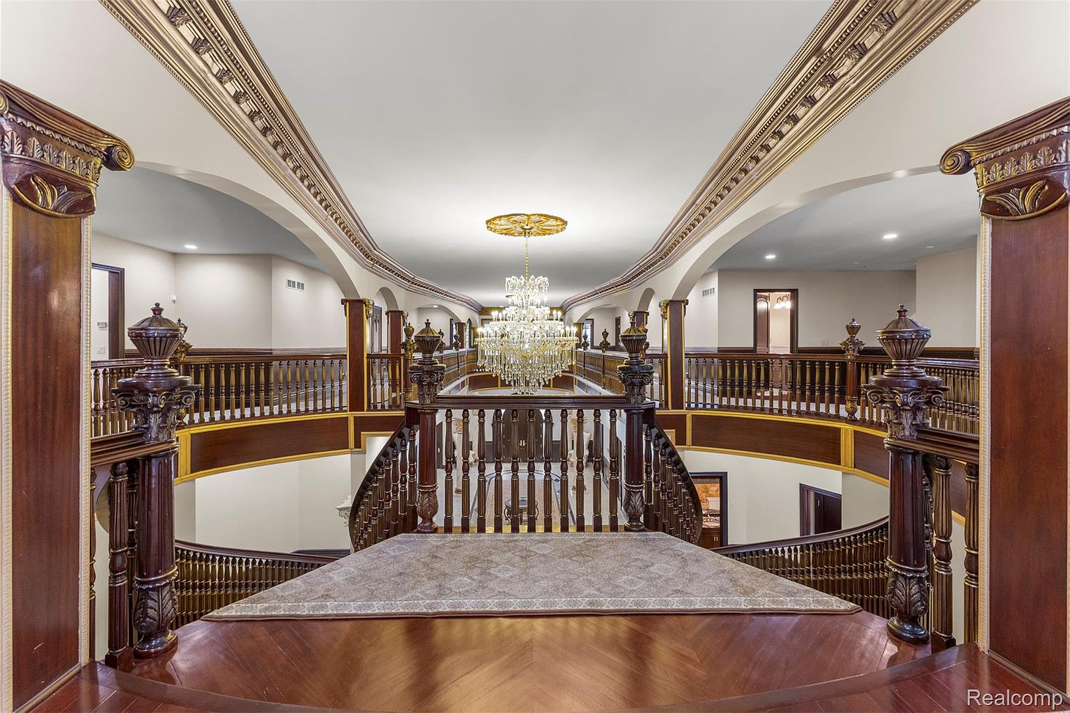 This grand interior shot showcases a luxurious hallway and staircase with ornate dark wood railings and columns. A large, multi-tiered crystal chandelier hangs from the ceiling, illuminating the space. The design features intricate architectural details and a sense of opulence, creating a sophisticated and elegant atmosphere.