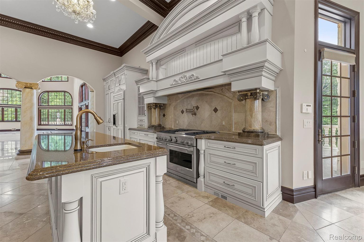 This is a luxurious kitchen featuring custom white cabinetry, granite countertops, and high-end stainless steel appliances. The focal point is a large decorative range hood with ornate detailing and column accents. The kitchen island includes a sink and faucet, and the flooring is a patterned tile, contributing to the room's elegant and upscale aesthetic.