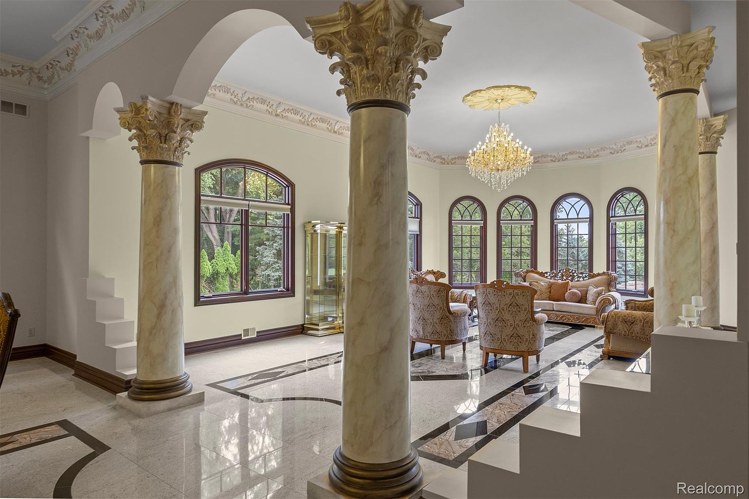 This grand living room features marble flooring, ornate columns, and a luxurious chandelier, creating an opulent and spacious atmosphere. Large arched windows provide ample natural light, highlighting the elegant furnishings and detailed architectural elements. The room exudes sophistication and timeless charm, perfect for entertaining or relaxing in style.