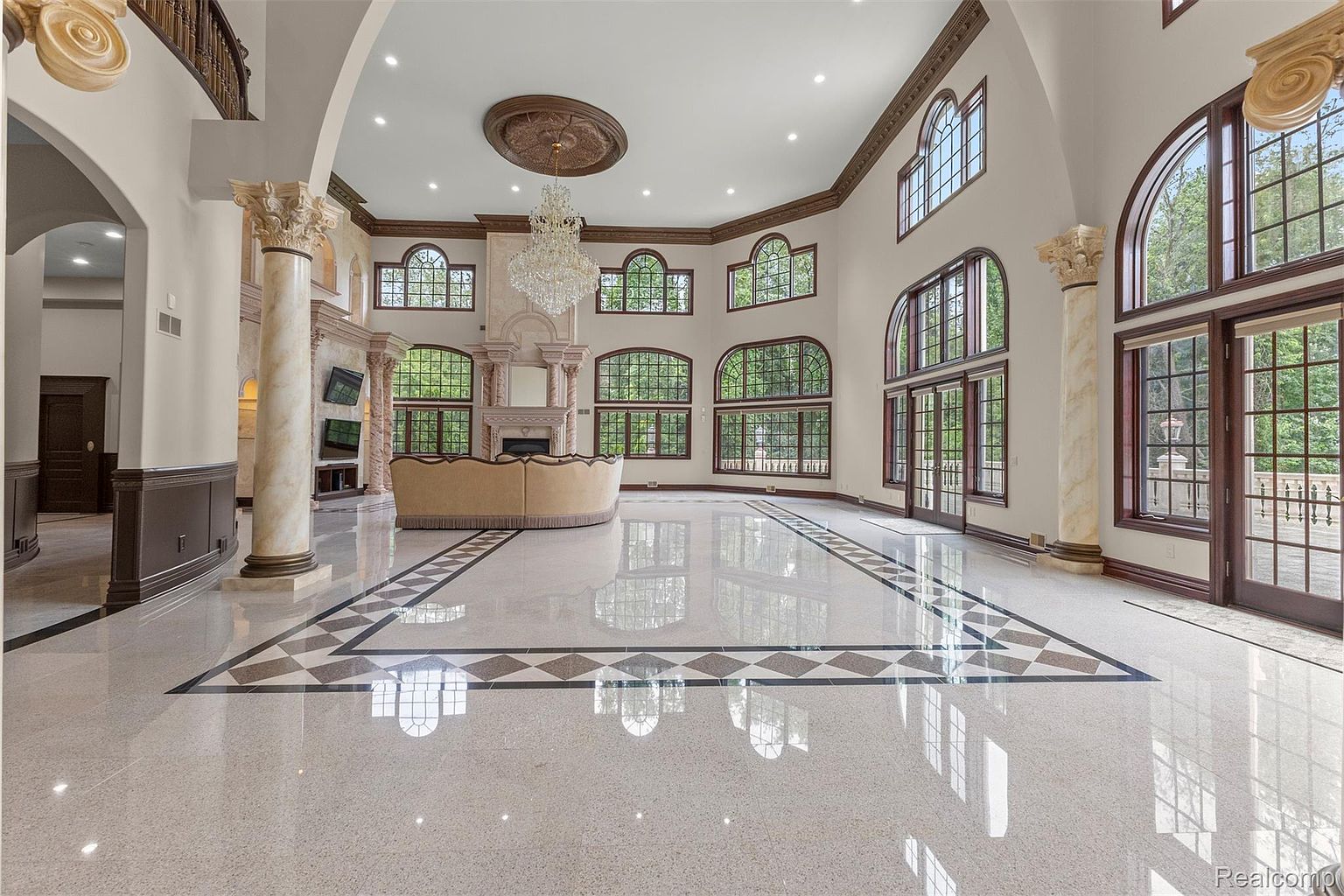 This is a grand living room featuring a high ceiling with an ornate ceiling medallion and a large crystal chandelier. The room is illuminated by natural light streaming through numerous arched windows. The flooring is a polished marble with a decorative border, and a curved sofa sits in the center of the room, facing a fireplace. The overall impression is one of luxury and spaciousness.