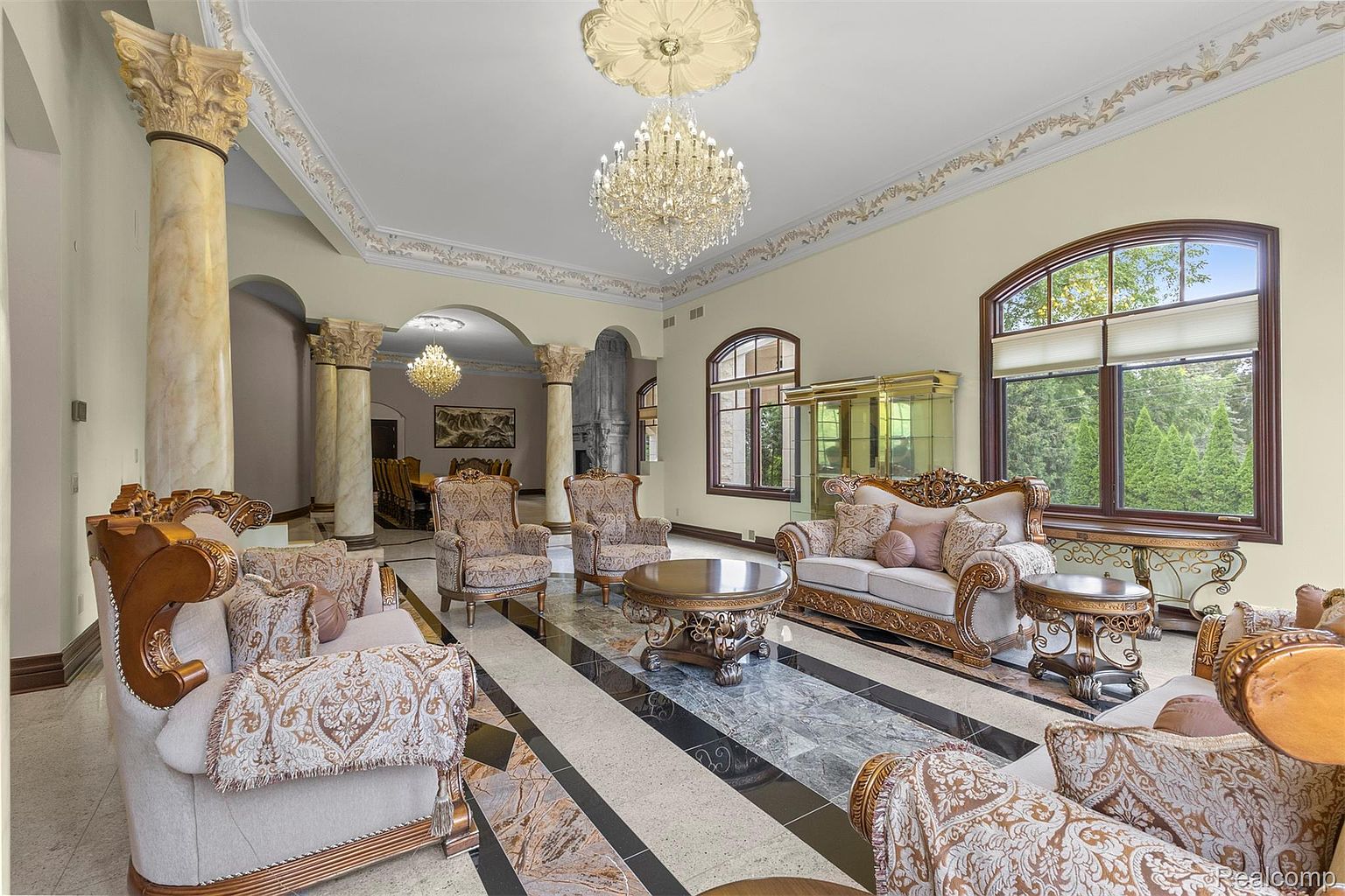 This is an opulent living room featuring ornate, gold-trimmed furniture with patterned upholstery. The room is adorned with marble columns, a large crystal chandelier, and decorative ceiling moldings. The flooring is a striking combination of black, white, and marble tiles, creating a luxurious and grand atmosphere.