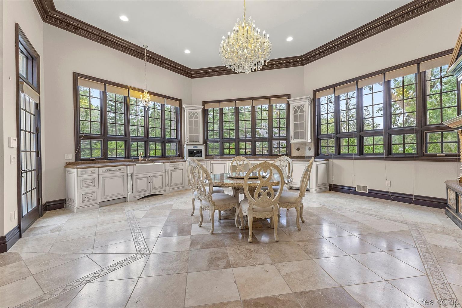 This elegant dining room features a round table with ornate chairs, set beneath a dazzling chandelier. Large windows surround the room, providing ample natural light and views of the outdoors. The room is finished with detailed trim work and tile flooring, creating a luxurious and inviting space.