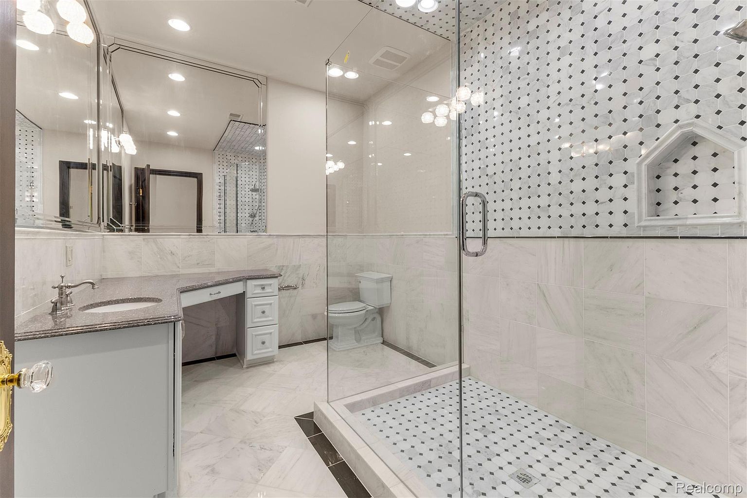 This is a primary bathroom featuring a vanity with a granite countertop, a large mirror, and a glass-enclosed shower. The shower has decorative tile and a built-in niche. The bathroom has a luxurious feel with its marble flooring and elegant fixtures.