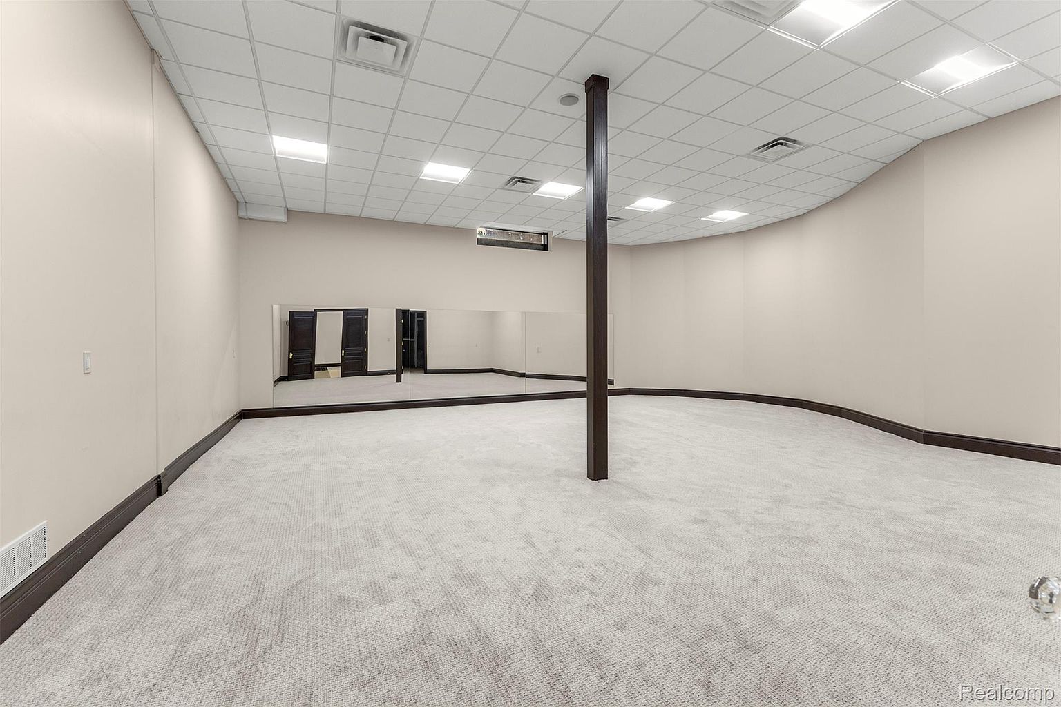 This is a spacious basement area featuring neutral-toned walls and light gray carpeting. A dark brown support beam stands prominently in the center of the room. A wall of mirrors is present, reflecting two dark-colored doors, suggesting a potential use as a dance studio or home gym.