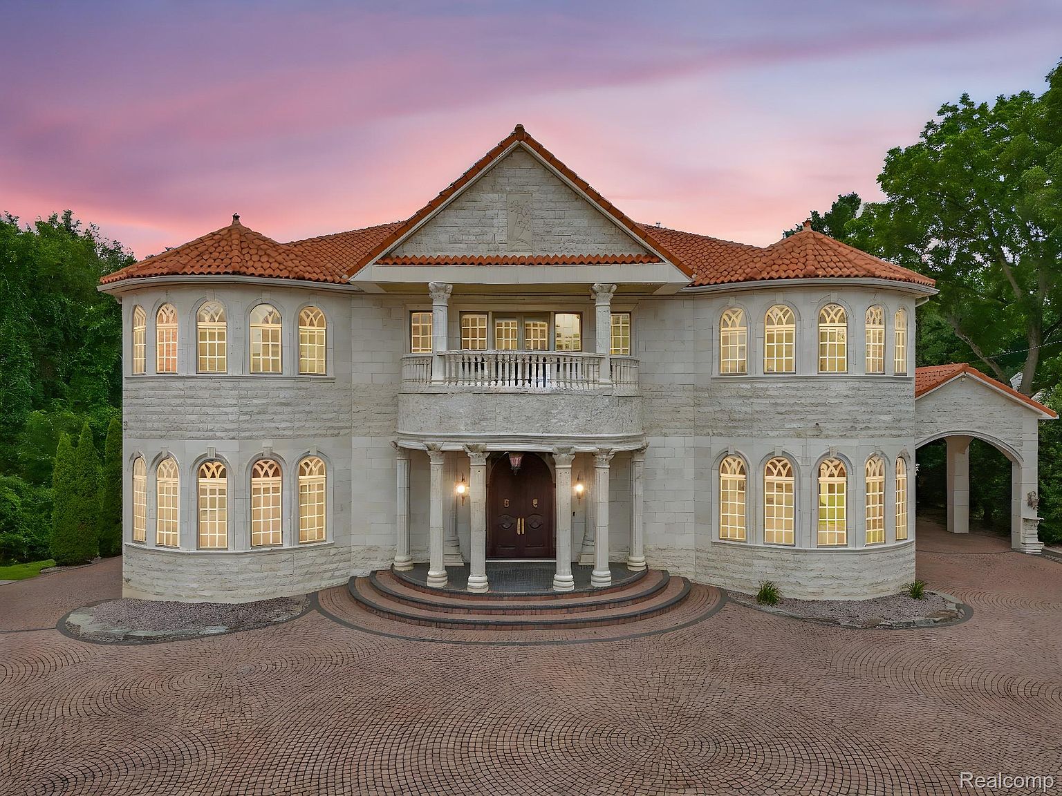 This is a front exterior view of a large, luxurious two-story house with a red tile roof and light-colored stone facade. The house features arched windows, a central balcony, and a grand entrance with columns and steps leading up to the front door. The driveway is paved with a circular brick pattern, and the sky is a beautiful gradient of pink and purple, suggesting either sunrise or sunset.