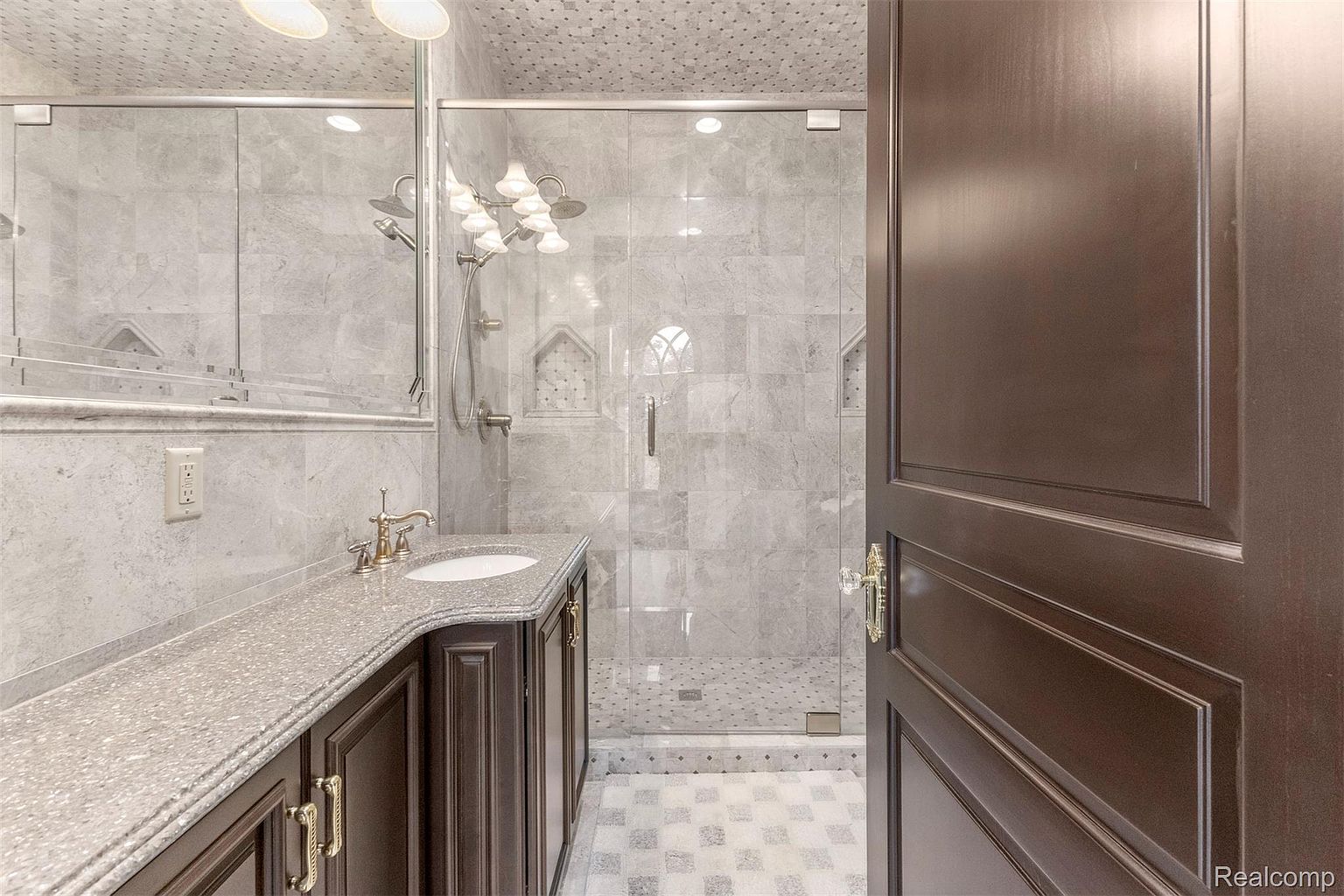 This is a well-appointed primary bathroom featuring a marble-tiled shower with glass enclosure, a vanity with a granite countertop, and dark wood cabinetry. The bathroom has a luxurious feel with its elegant fixtures, patterned floor tiles, and a shower with dual showerheads. The door is partially open, inviting the viewer into the space.