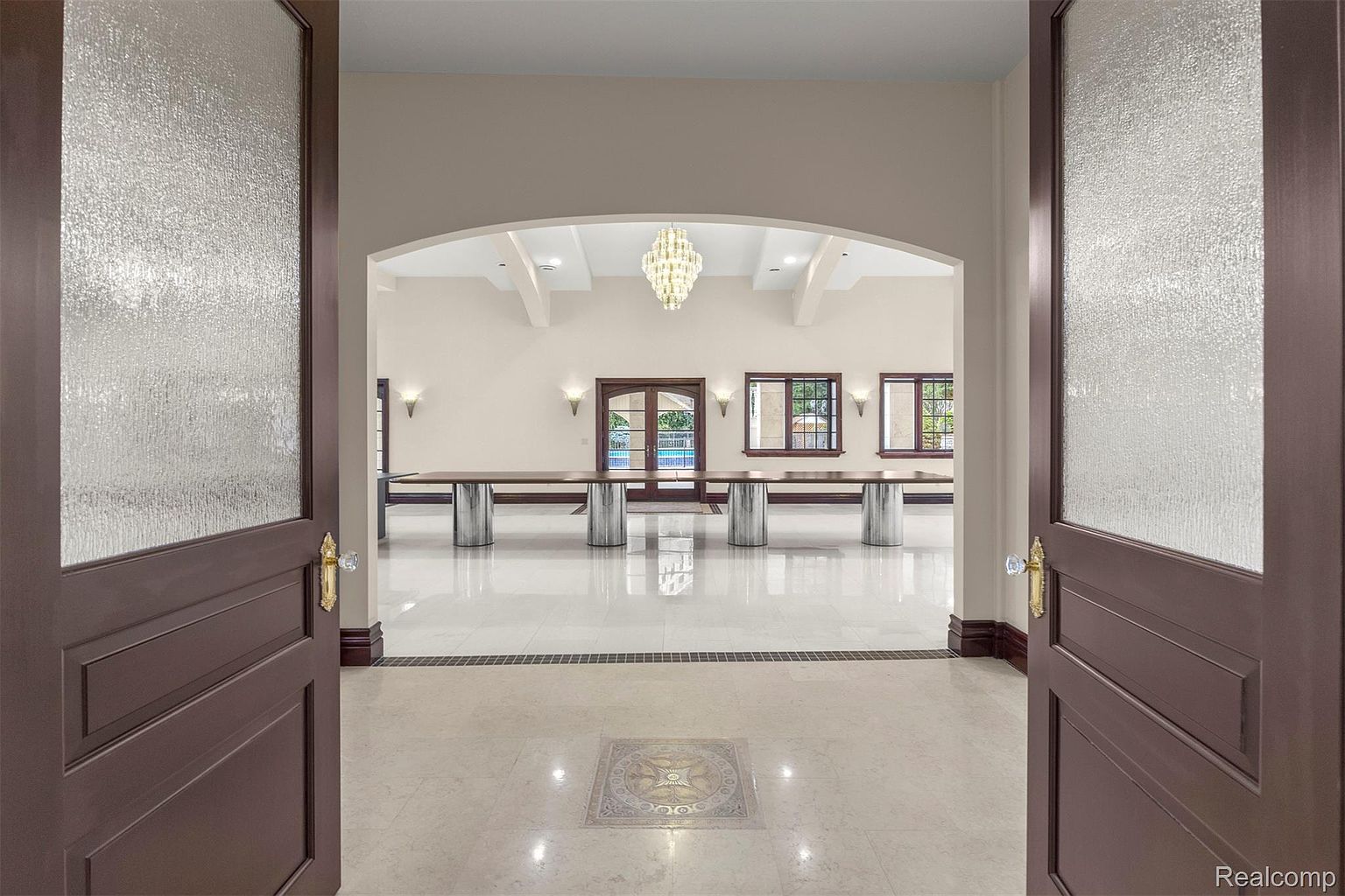 This interior shot showcases a grand dining room, viewed through an open doorway. The room features a long dining table with unique cylindrical supports, a decorative chandelier, and large windows that provide natural light. The polished tile flooring and neutral wall colors contribute to an elegant and spacious atmosphere, perfect for entertaining.