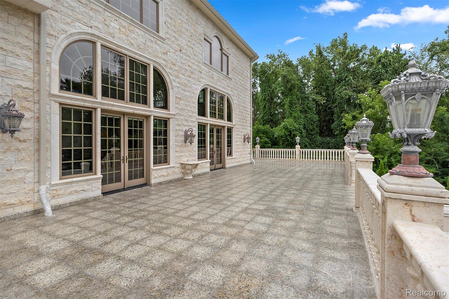 This image showcases a spacious patio or balcony area of a luxurious home. The area features a patterned stone floor, a decorative stone railing with ornate lampposts, and large arched windows and doors leading into the house. The exterior of the house is clad in light-colored stone, creating an elegant and upscale impression.
