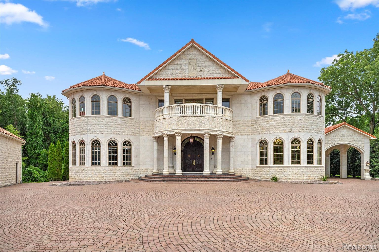 This is a grand front view of a luxurious, two-story mansion. The house features a light-colored stone facade, a red tile roof, and a prominent balcony supported by columns. A circular brick driveway leads up to the entrance, enhancing the property's curb appeal and suggesting a high-end, elegant lifestyle.