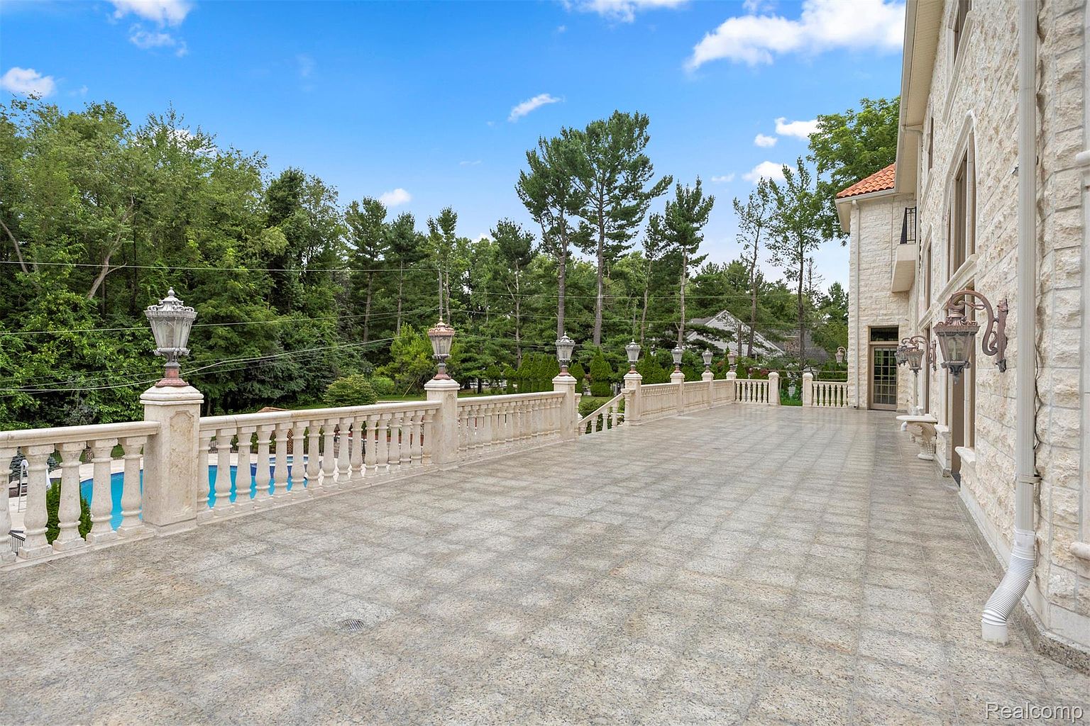 This image showcases a spacious patio or balcony area of a luxurious home, featuring a decorative balustrade with ornate lighting fixtures. The flooring is patterned tile, and the view overlooks lush greenery and a glimpse of a pool. The architectural style suggests high-end construction and design.