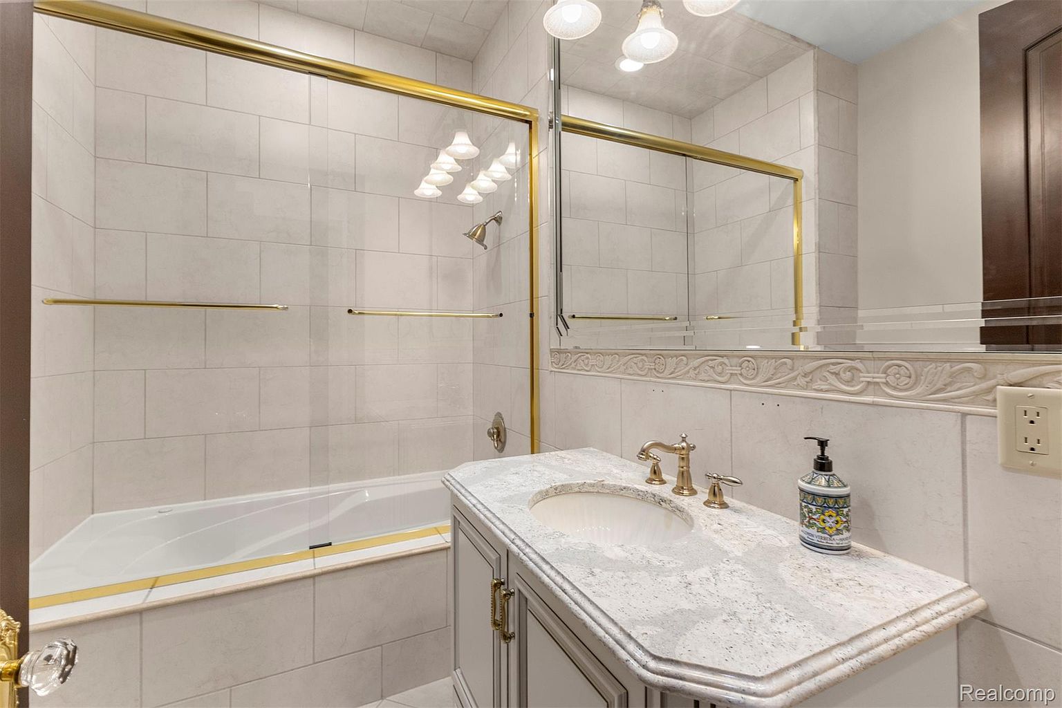 This is a primary bathroom featuring a bathtub with a gold trim, a glass shower door with gold trim, and a vanity with a white countertop. The walls are tiled in a neutral color, and the overall style is classic and elegant. The bathroom appears clean and well-maintained.