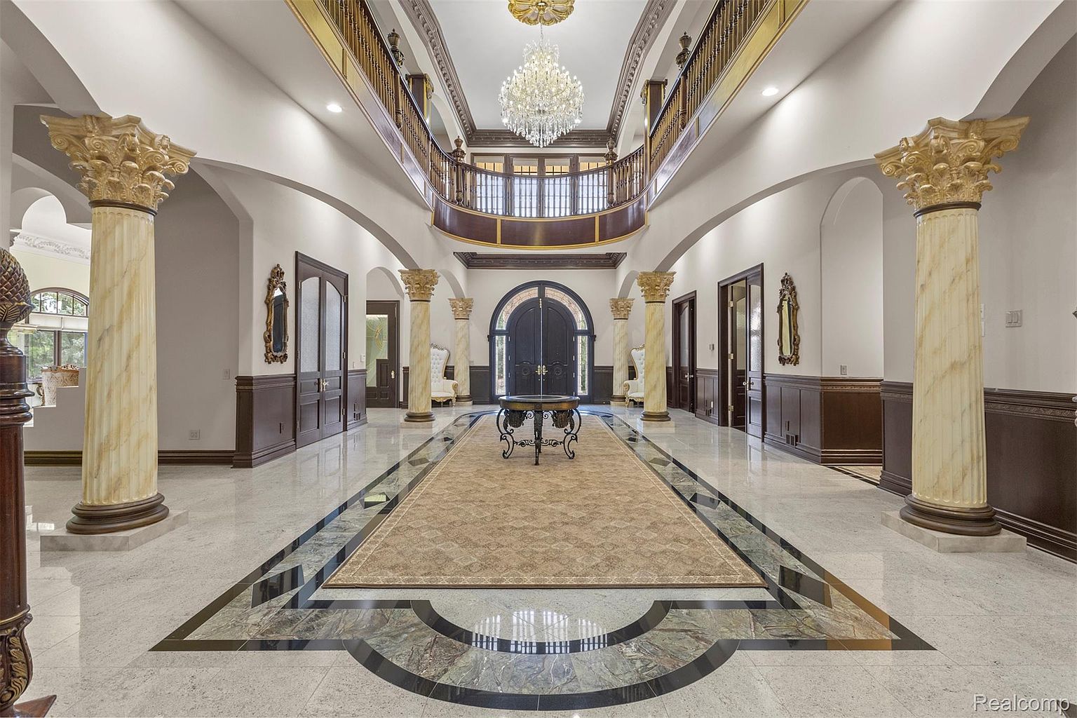 This grand foyer features marble flooring with a decorative inlay, flanked by large columns with ornate capitals. A long runner rug leads to a set of double doors, with a table in the center. A second-story balcony overlooks the foyer, illuminated by a large chandelier, creating a luxurious and impressive entrance.